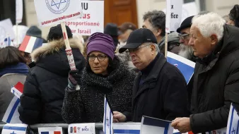 الصورة تظهر مجموعة من الأشخاص في تجمع أو مظاهرة. يحملون لافتات وأعلام، من بينها العلم الإسرائيلي، ويرتدون ملابس شتوية. يبدو أن المتظاهرين يعبرون عن آرائهم حول قضية معينة تتعلق بإسرائيل. المشهد يعكس تفاعل الناس مع قضايا سياسية واجتماعية.