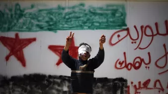 تظهر الصورة صبيًا صغيرًا يرفع يديه بإشارات النصر، بينما يغطّي عينيه برباط أبيض. خلفه جدار مطلي عليه رسم يمثل علم سوريا، مع كلمات مكتوبة بالعربية تظهر اعتراضًا أو دعماً لموضوع معين. تعكس الصورة مشاعر الأمل والعزيمة في وجه الصعوبات.