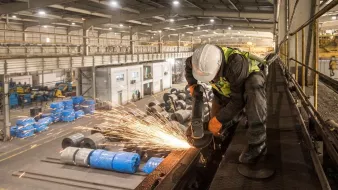 تظهر في الصورة عاملاً يرتدي خوذة وسماعات أذن، بينما يقوم بعملية قطع أو طحن المعادن في مصنع. تتطاير الشرر أثناء عملية العمل، مما يضيف إلى الحركة والديناميكية في المكان. خلف العامل، يُمكن رؤية مساحة واسعة من المصنع تضم معدات وعجلات مغطاة بأقمشة زرقاء، مما يشير إلى بيئة صناعية نشطة. الإضاءة في الصورة جيدة، مما يبرز تفاصيل العمل والبيئة المحيطة.