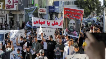 الصورة تُظهر مجموعة من المتظاهرين في شارع يجمعهم الاحتجاج، حيث يحملون لافتات كبيرة تحمل شعارات ومطالب تعبر عن قضايا معينة. يظهر في الصورة عدد من الرجال والنساء والأطفال، وبعضهم يرتدي القمصان بتصاميم مختلفة. يبدو الحشد نشطاً ومتفاعلاً، واللافتات مكتوبة باللغة العربية، مما يدل على أن التظاهر يتناول قضايا محلية. الجو يبدو حماسياً مع وجود بعض السيارات في الخلفية.