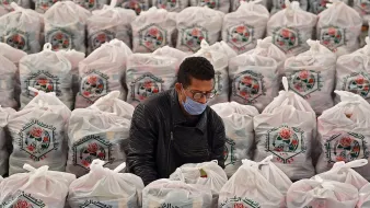 الصورة تظهر رجلًا يرتدي قناع وجه وهو يجلس بين صفوف من أكياس كبيرة، حيث تحتوي هذه الأكياس على مواد غذائية أو مستلزمات معينة. الأكياس تحمل شعارًا معينًا ويبدو أنها منظمة أو جمعية ما. الرجل يبدو مهتمًا بما يقوم به، ويعمل على ترتيب الأكياس أو تجهيزها. الخلفية مليئة بالأكياس بشكل منظم، مما يشير إلى نشاط يتعلق بتوزيع المساعدات أو العمل الخيري.