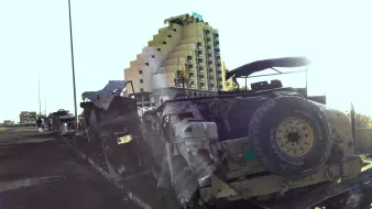 الصورة تظهر مجموعة من المركبات العسكرية المدمرة أو المهجورة على جانب الطريق. في الخلفية، يوجد مبنى شاهق يبدو مهجوراً، ويتميز بتصميمه الفريد. تضيء الشمس الصورة من جهة واحدة، مما يبرز تفاصيل المركبات والمبنى. الجو العام يوحي بالتخلي أو الفوضى، ويعكس مشاهد قد تكون ناتجة عن حالة نزاع أو حرب.
