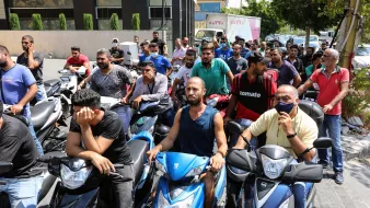 تظهر الصورة مجموعة من الأشخاص يتجمعون أمام مركز ما، حيث يبدو أنهم في انتظار الحصول على شيء ما. معظمهم يركبون دراجات نارية، ويبدو عليهم التوتر والقلق. بعضهم يظهر علامات الإحباط، بينما آخرون يتحدثون مع بعضهم. الخلفية تحتوي على مباني وأشجار، مما يشير إلى أن المشهد في منطقة حضرية.