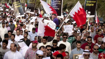 الصورة تظهر تجمعًا حاشدًا لمتظاهرين في البحرين يحملون لافتات وأعلام البلاد. يتواجد الأشخاص في مكان مفتوح، ويسود الجو شعور بالتضامن والمطالبة بالتغيير. اللافتات تتضمن مطالب للديمقراطية ورفض السياسات القمعية. يرتدي المتظاهرون ملابس تقليدية وطنية، ويبدو أن الحشد متنوع من حيث الأعمار.
