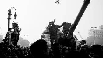 تظهر الصورة مشهداً من مظاهرة أو حدث احتجاجي، حيث يوجد عدد كبير من الناس يتجمعون حول دبابة. أحد الجنود يقف فوق الدبابة ويرفع يدَه كما لو كان يحمل شيئاً أو يجذب الانتباه. الأجواء تشير إلى توتر أو حالة من الحماس بين الحشود. الصورة تُظهر تبايناً كبيراً بين الأضواء والظلال، مما يضفي عليها طابعاً دراماتيكياً.