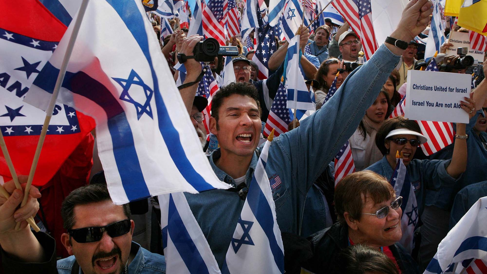 The image depicts a large gathering of people holding flags, primarily Israeli and American, as they participate in a rally or demonstration. The crowd appears to be energetic and passionate, with individuals expressing enthusiasm—some are cheering, while others are waving their flags. The setting seems to be outdoors, likely in a public space, and the atmosphere conveys a sense of unity and support for a cause related to Israel.