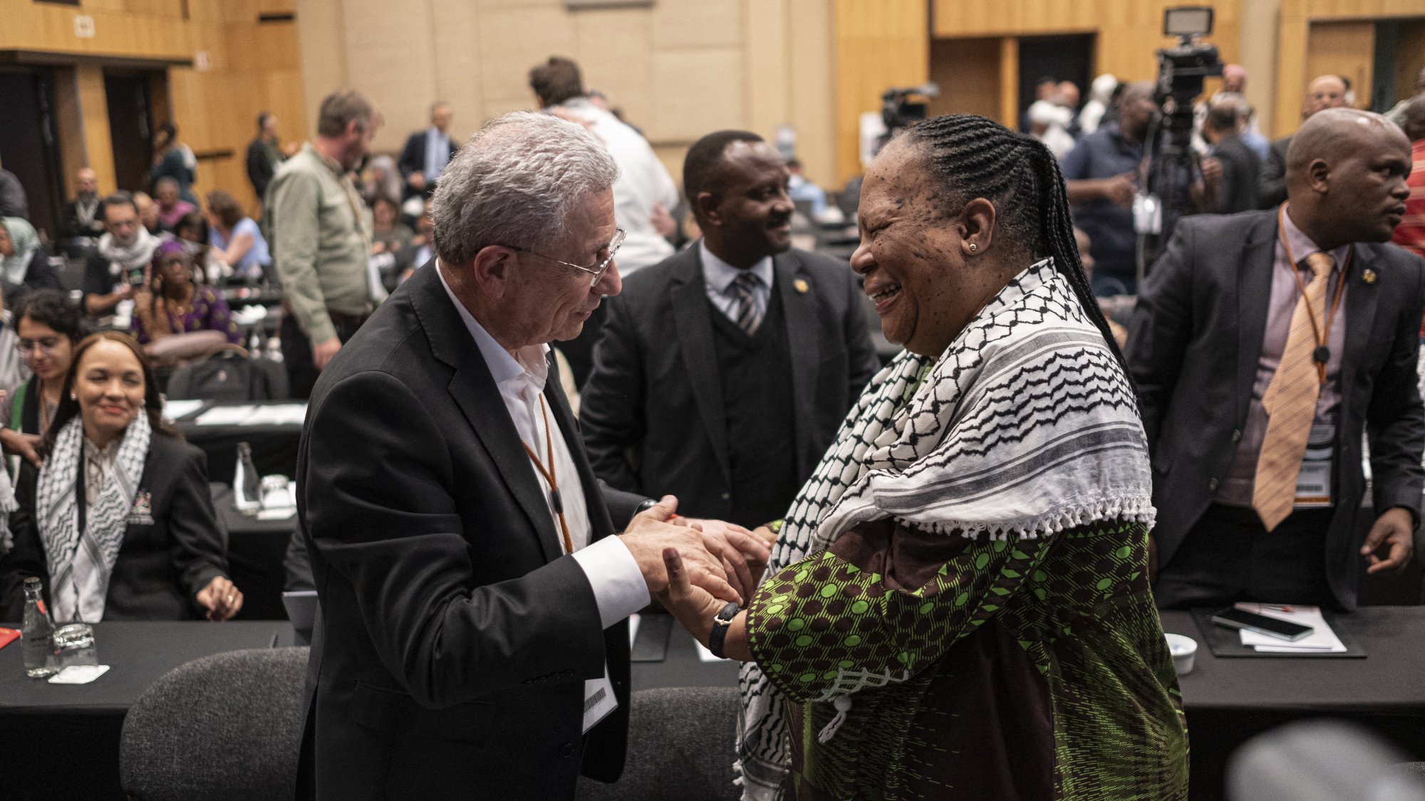 The image depicts a moment of interaction between two individuals at a conference or event. Both people are smiling and engaged in a handshake, suggesting a friendly and positive exchange. The person on the left is wearing a suit, while the individual on the right is dressed in traditional or cultural attire with a colorful shawl. In the background, there are other attendees, some seated and others standing, indicating a busy atmosphere typical of a conference. The setting appears to be indoors, likely within a large hall or meeting room.
