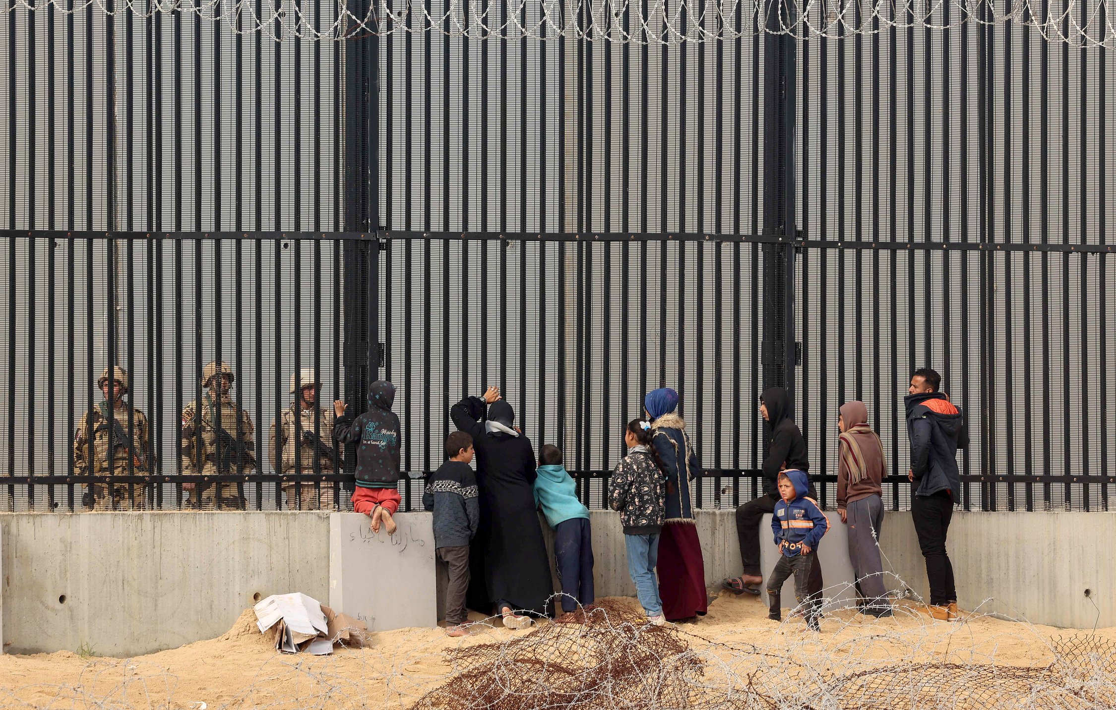 The image depicts a group of people, including women and children, standing in front of a tall, barred fence. On the other side of the fence are soldiers or guards, who appear to be watching the group. The scene conveys a sense of separation and tension, with barbed wire in the foreground emphasizing the barriers between the two sides. The individuals on the left seem to be engaging with the guards, possibly seeking assistance or communication. The overall atmosphere suggests a complex and challenging situation, likely related to migration or security issues.