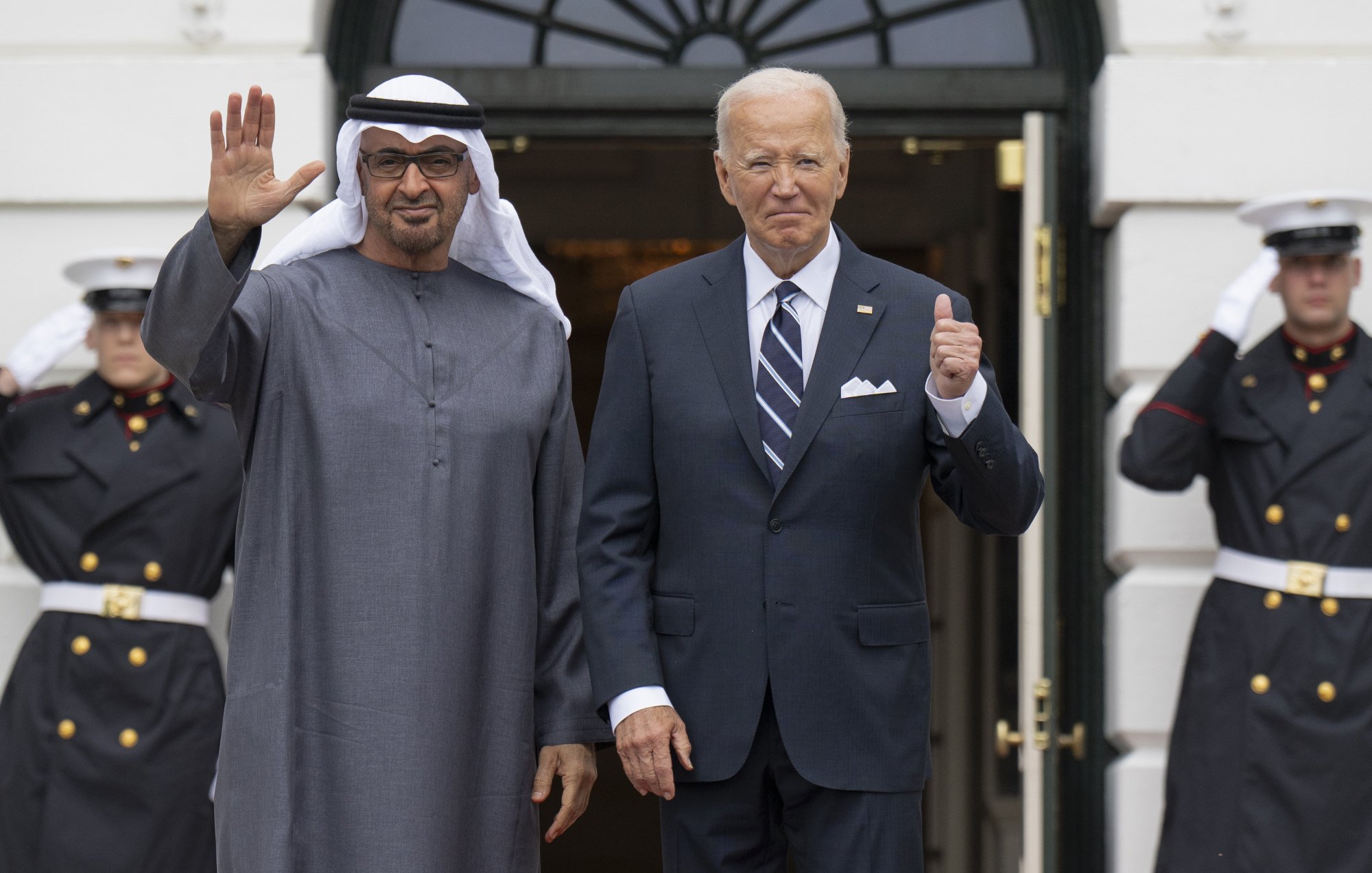 The image shows two men standing at the entrance of a building, likely the White House. One man is dressed in traditional attire with a white headscarf, while the other is in a formal suit. They are both smiling and posing for the camera, with one man waving and the other giving a thumbs-up. In the background, there are uniformed guards standing at attention. The scene conveys a sense of diplomacy and cordiality between the two leaders.