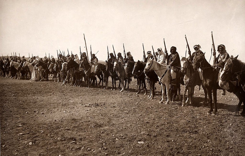 The image depicts a line of mounted warriors or soldiers, likely from a historical context. They are on horseback, holding spears or similar weapons, and are dressed in traditional clothing that suggests their cultural identity. The setting appears to be an open, dry landscape, possibly a desert or arid region. The overall sepia tone gives the photograph an aged appearance, typical of early 20th-century photography. The formation and attire indicate that it could represent a military or ceremonial gathering.