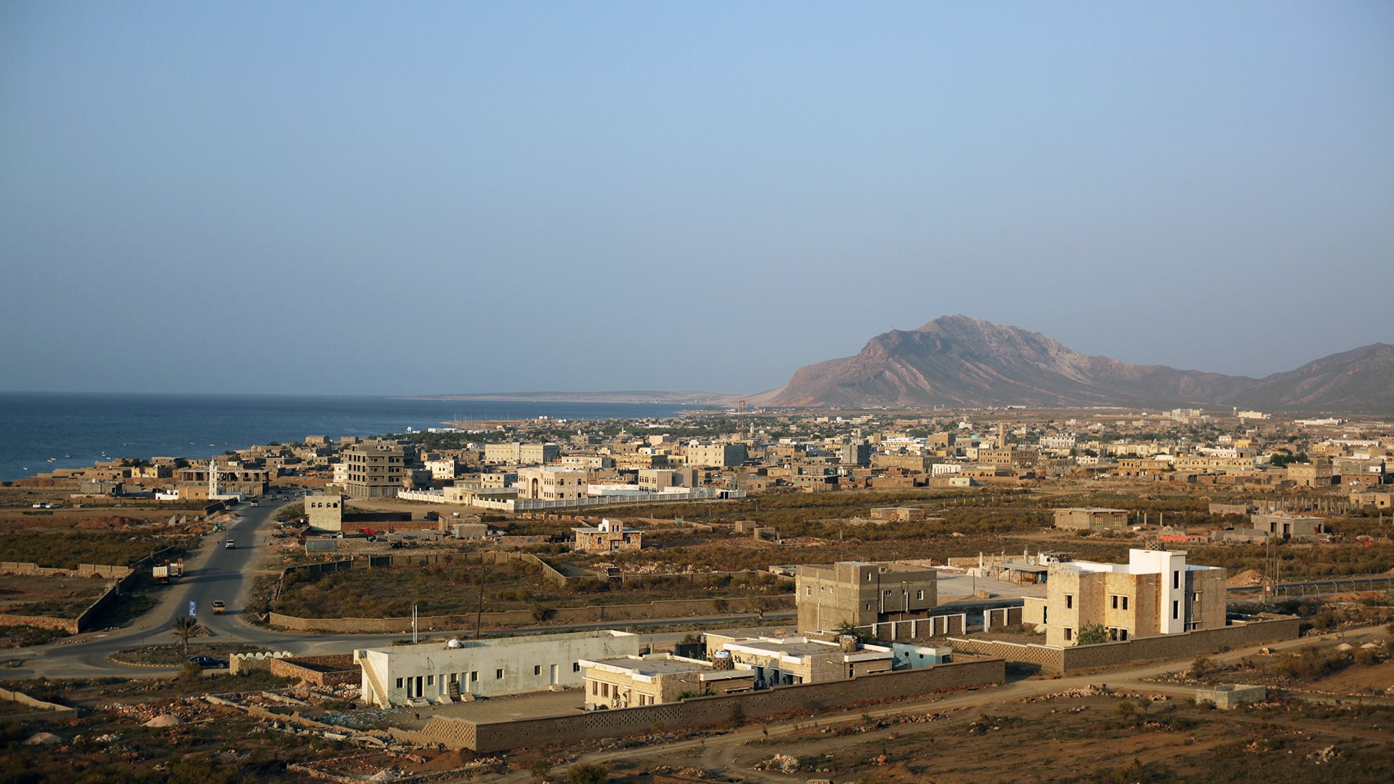 The image depicts a coastal landscape with a view of a small town or village near the ocean. In the foreground, there are a few buildings, some of which are modern, while others appear more traditional. The road winds through the landscape, leading towards the sea. In the background, a mountain rises against a clear sky, adding to the natural beauty of the scene. The overall atmosphere suggests a quiet, possibly rural environment with a mix of natural and man-made elements.