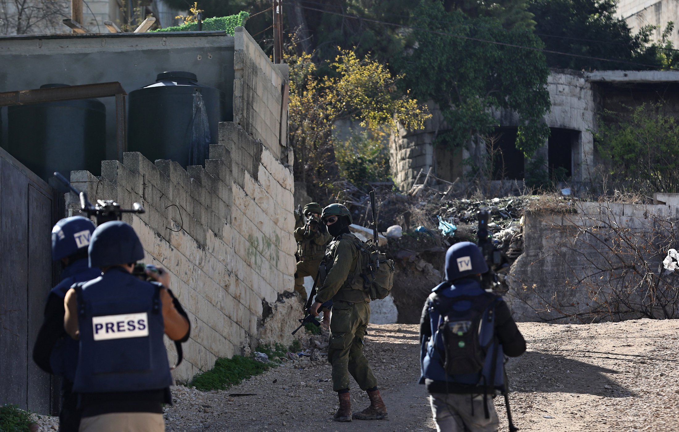The image depicts a scene in an urban environment, likely involving a military or conflict situation. In the foreground, there are three individuals wearing vests labeled "PRESS," indicating they are journalists or reporters. They appear to be observing or documenting the situation. In the background, a soldier in military gear is positioned, holding a weapon and looking in the direction of the journalists. The surroundings include a wall, some buildings, and debris, suggesting the area may be experiencing instability or conflict.