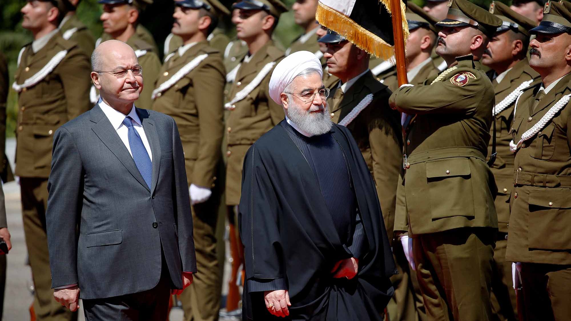 The image shows two men walking in a formal setting, likely during a ceremonial event. One man is dressed in a suit and tie, while the other is wearing a traditional black robe and a white turban. They are surrounded by a group of uniformed soldiers standing at attention, indicating a military or state occasion. The atmosphere appears formal and significant, suggesting a diplomatic meeting or official visit.