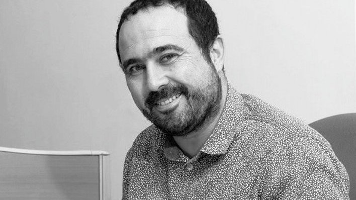 The image shows a man sitting and smiling. He has a short beard and is wearing a spotted button-up shirt. The background appears to be a plain wall and a piece of furniture, suggesting a professional or office setting. The overall tone of the image is friendly and approachable.