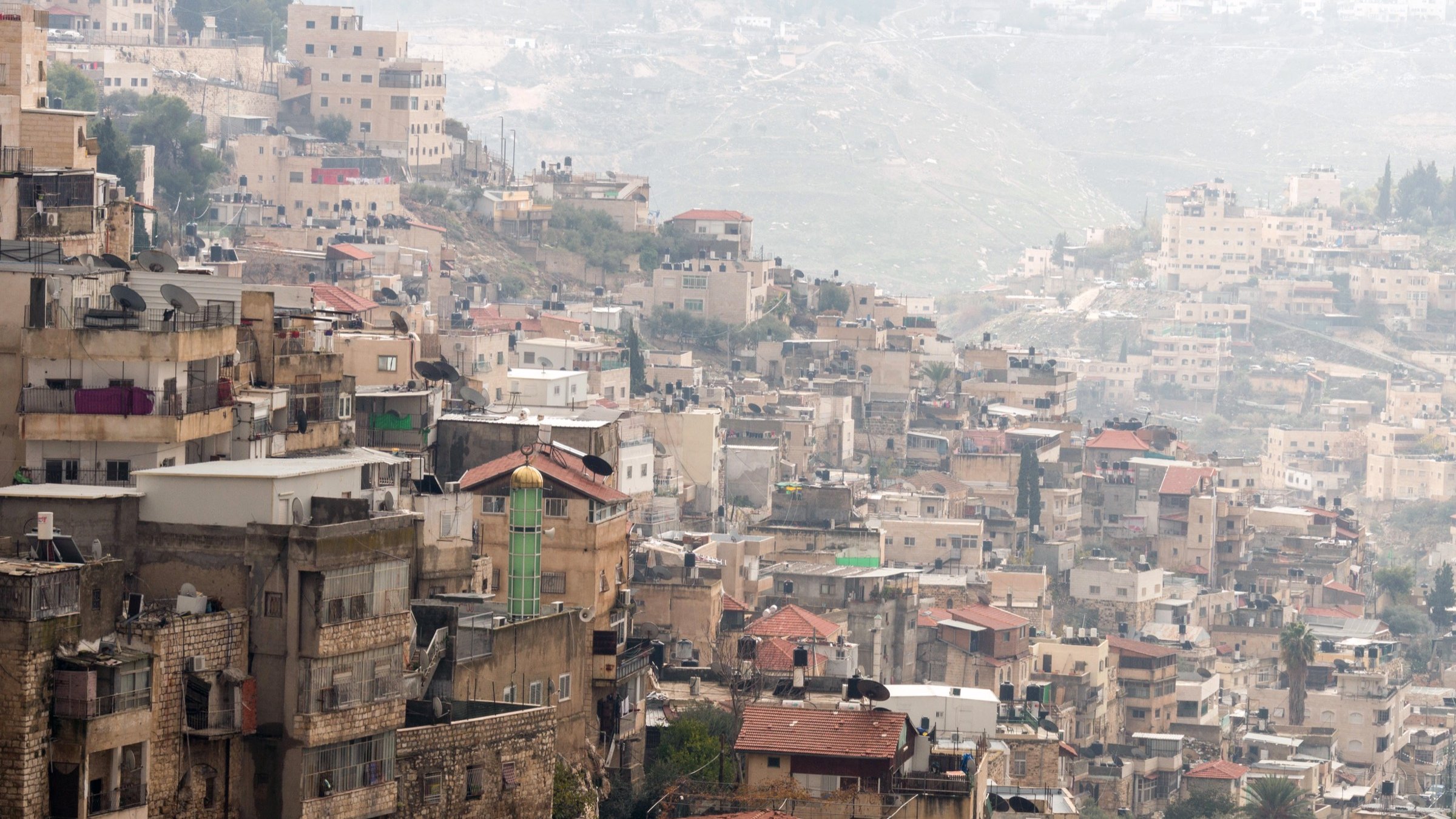 The image shows a densely populated hillside area with a mix of residential buildings. The structures vary in height and architectural style, with many appearing stacked closely together. Some roofs are visible, displaying a range of materials, from tiles to concrete. In the background, more houses and buildings can be seen on the sloping terrain, suggesting a continuation of urban development. The overall scene has a somewhat hazy atmosphere, possibly due to distance or weather conditions.