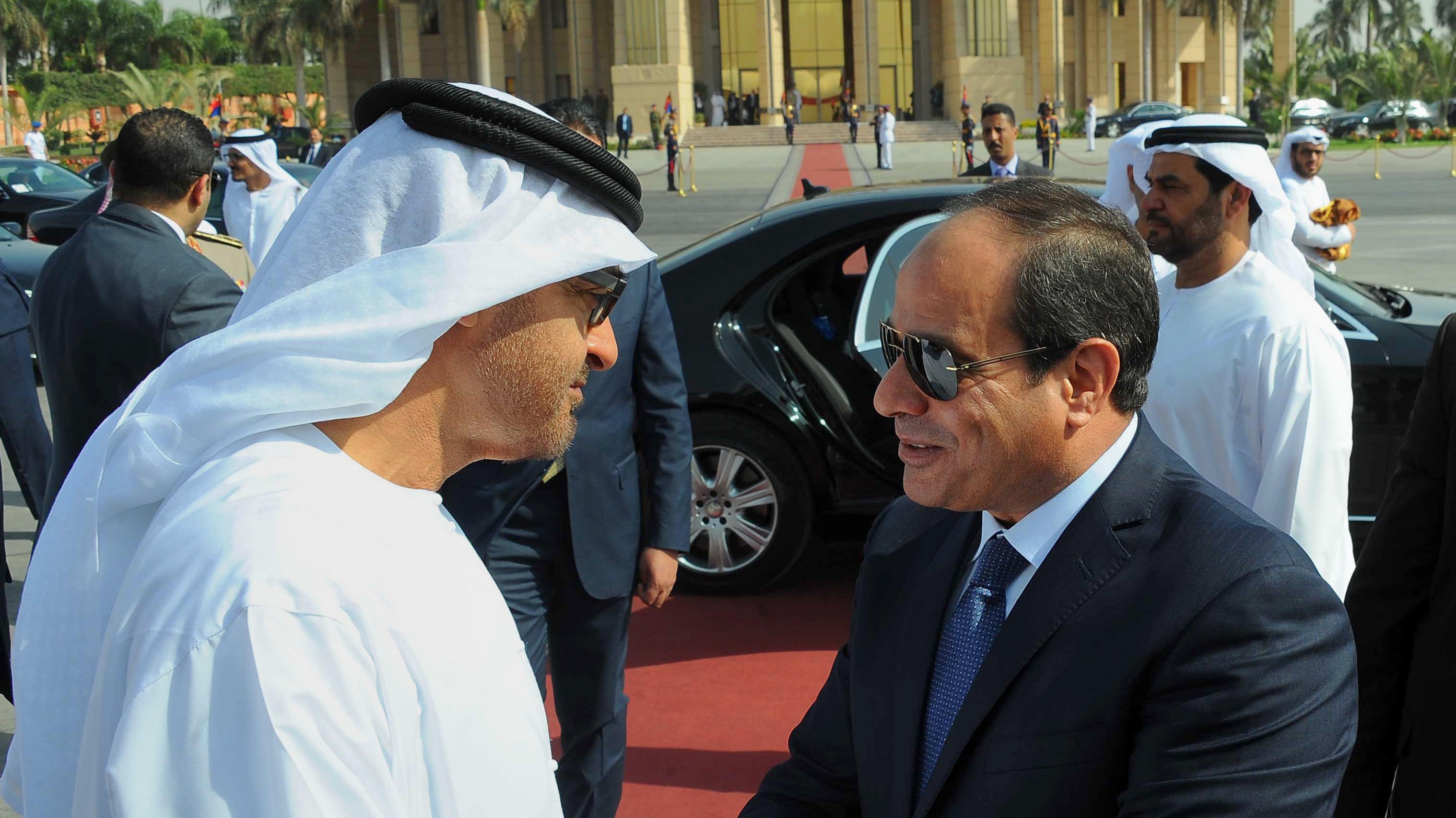 The image depicts two men in formal attire engaged in a handshake, suggesting a cordial greeting. One man is dressed in traditional Emirati clothing, including a white kandura and headscarf, while the other is wearing a dark suit and tie. They appear to be in an official setting, with a luxurious car and a group of well-dressed individuals in the background. The atmosphere indicates a diplomatic or state visit, emphasizing cooperation and respect between the two figures.