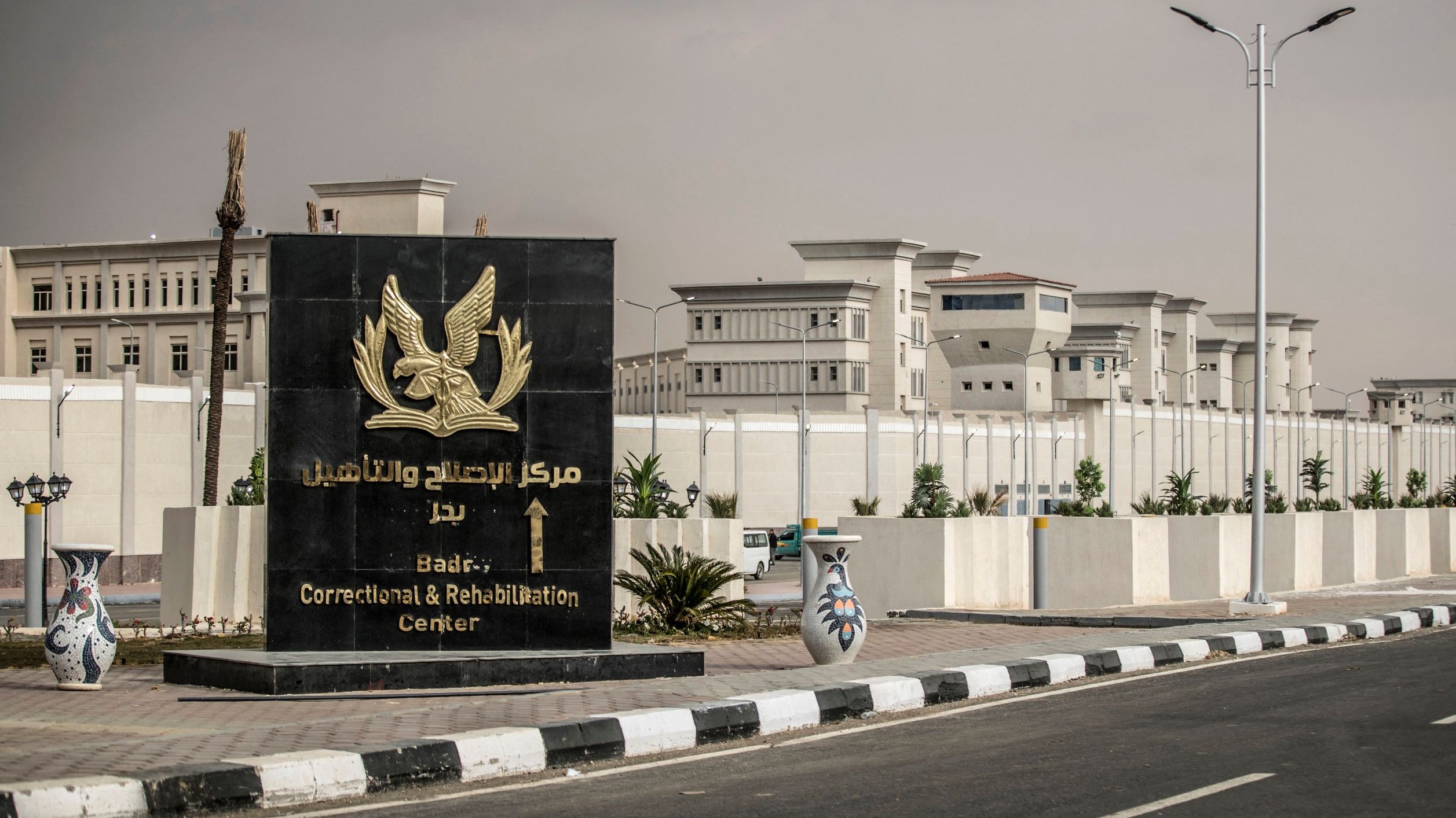 The image depicts a large sign featuring an eagle emblem, which is located in front of a correctional and rehabilitation center. The sign is written in both Arabic and English, indicating the name of the facility, which is "Badry Correctional and Rehabilitation Center." The center itself is visible in the background, showcasing a well-maintained structure surrounded by a tall wall and clear skies. There are decorative elements at the base of the sign, including ornamental vases, and the area is lined with street lamps.