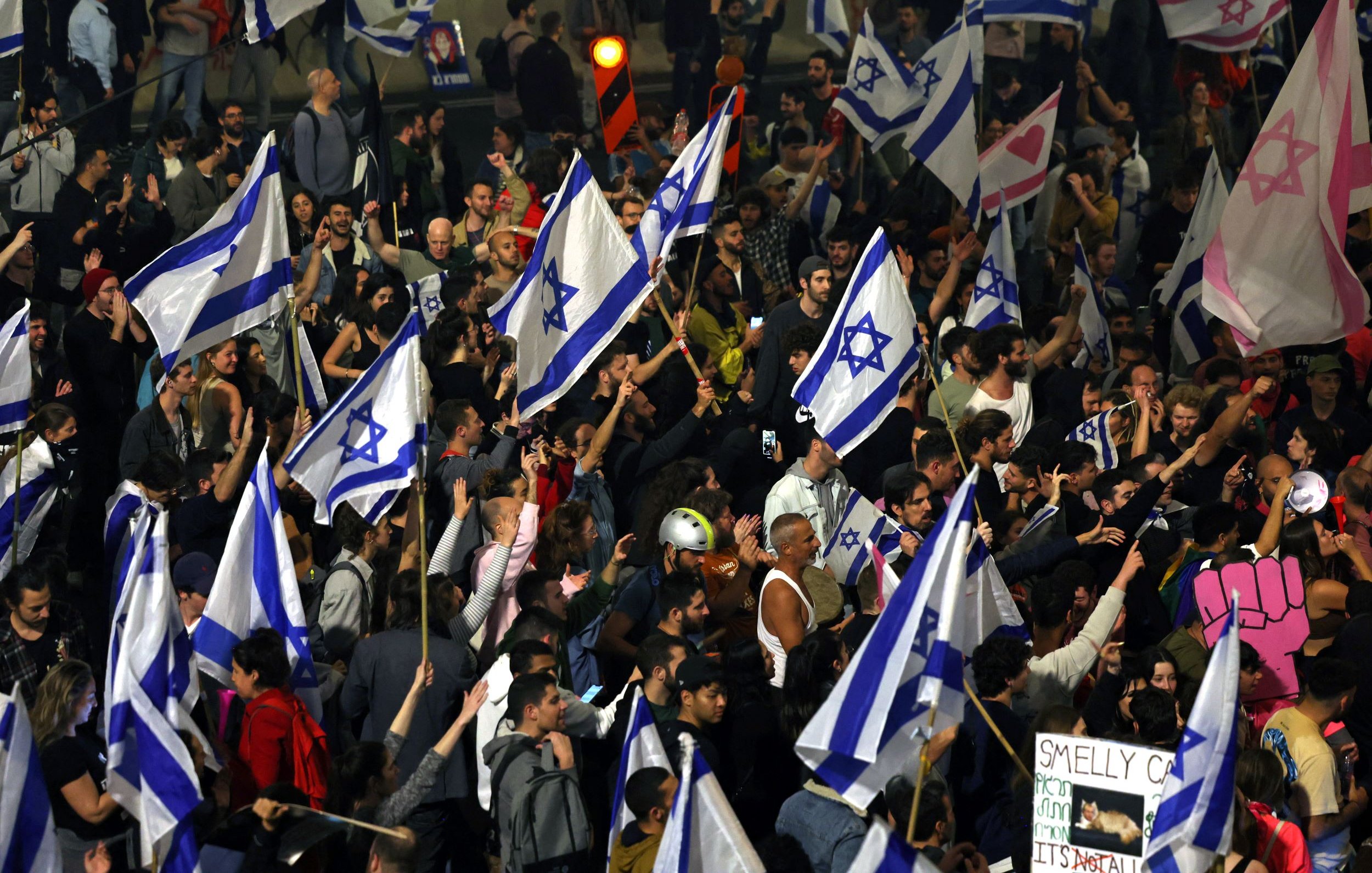 The image depicts a large crowd of people gathered together, many of whom are holding Israeli flags. The atmosphere appears vibrant and energetic, with a mix of participants visible, some raising their hands and signs. Among the signs, one reads "SMELLY C**" in a playful manner. The crowd suggests a protest or demonstration, characterized by a sense of unity and purpose, illuminated by city lights in the background.