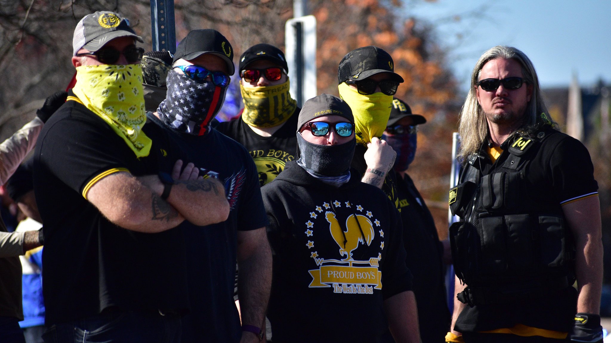 The image depicts a group of people standing together, wearing clothing that features dark colors and some yellow accents. Many of them have their faces covered with bandanas or masks, and they are wearing sunglasses. Their expressions and postures suggest a sense of solidarity or confrontation. The group is likely gathered for a specific event or rally, as indicated by their coordinated appearance. In the background, there are trees and other people visible, suggesting an outdoor setting.