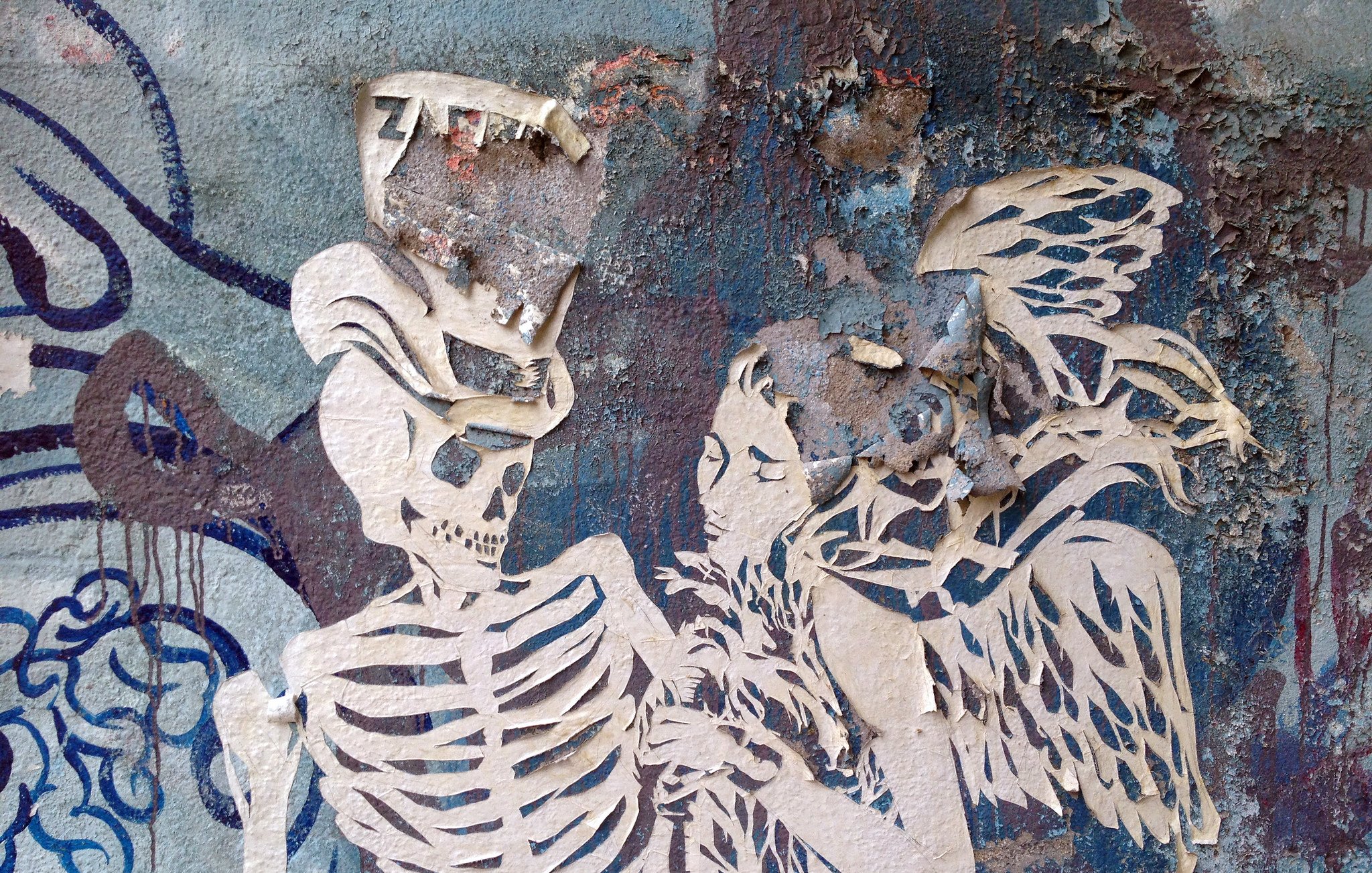 The image features a graffiti-style artwork on a textured wall. It depicts a skeletal figure, a skeleton, in an intimate pose with a woman who seems to have angelic features, including wings. The artwork appears to be layered, with parts of the paper or paint peeling away, adding a weathered and distressed look. The background consists of blue and gray tones, enhancing the overall atmosphere of the piece. The combination of the skeleton and the woman creates a striking contrast, possibly symbolizing themes of life and death, or love and loss.