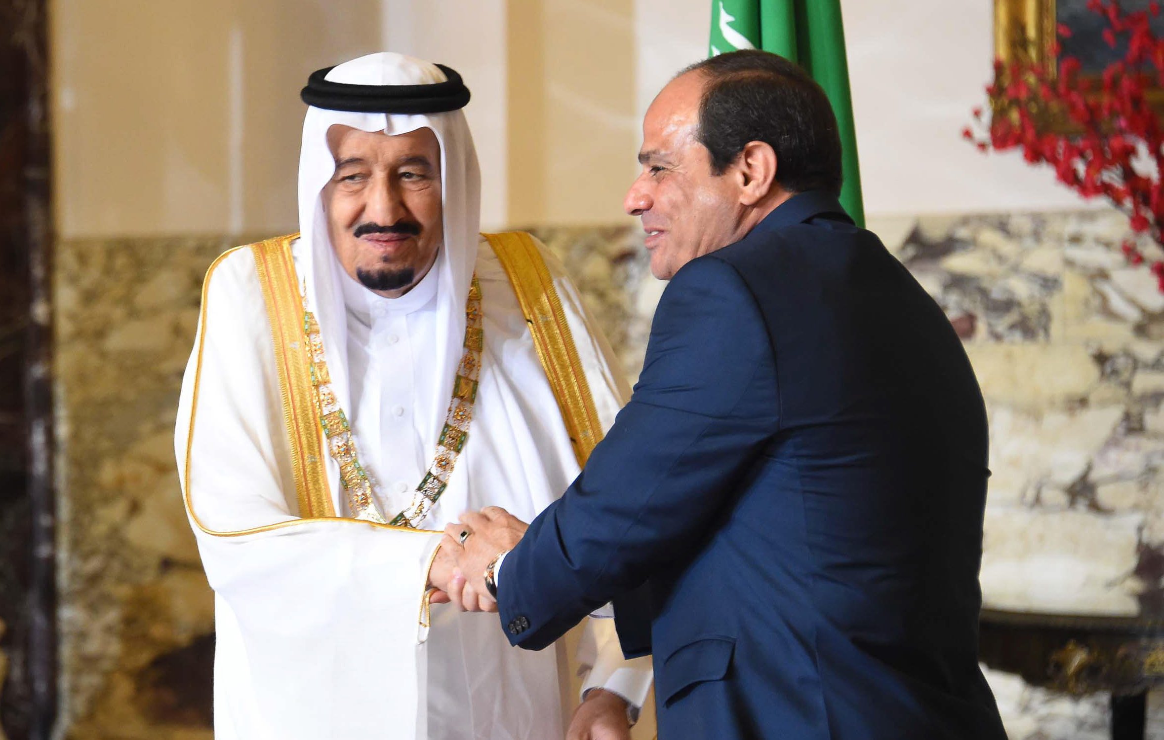 The image depicts two men in formal attire shaking hands. One man is wearing traditional Saudi clothing, including a white thobe and a black and white headscarf, adorned with a gold sash. The second man is dressed in a dark suit. They appear to be engaged in a cordial greeting, likely in a diplomatic or official context. In the background, there are hints of decor that suggest an elegant setting, possibly within a government building. One of the men is also positioned near a flag, indicating a connection to national representation.