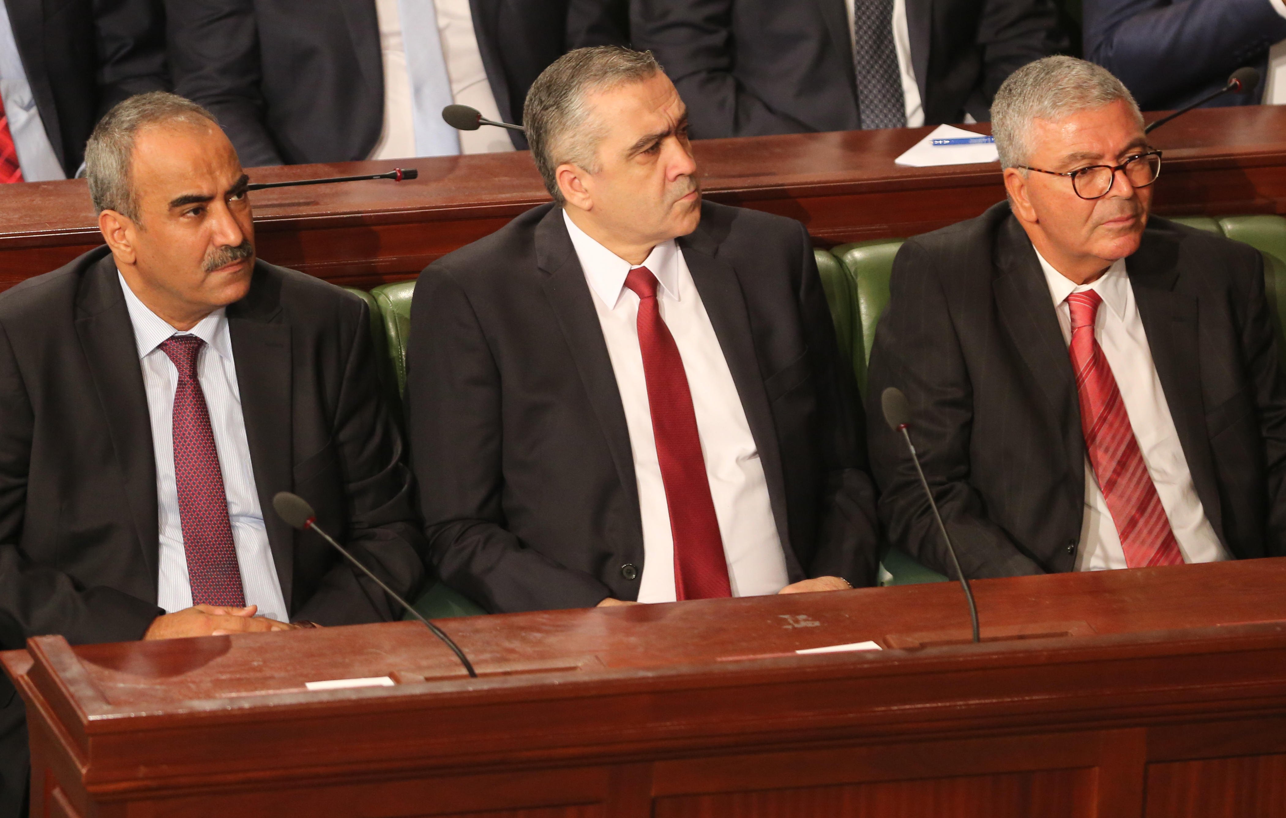 The image shows three men sitting side by side in a formal setting, likely within a legislative or governmental assembly. They are dressed in business suits, with two wearing ties—one in red and the other in a lighter color. The background suggests a gathering or meeting, with a group of people seated behind them. The scene conveys a sense of seriousness and official duty.