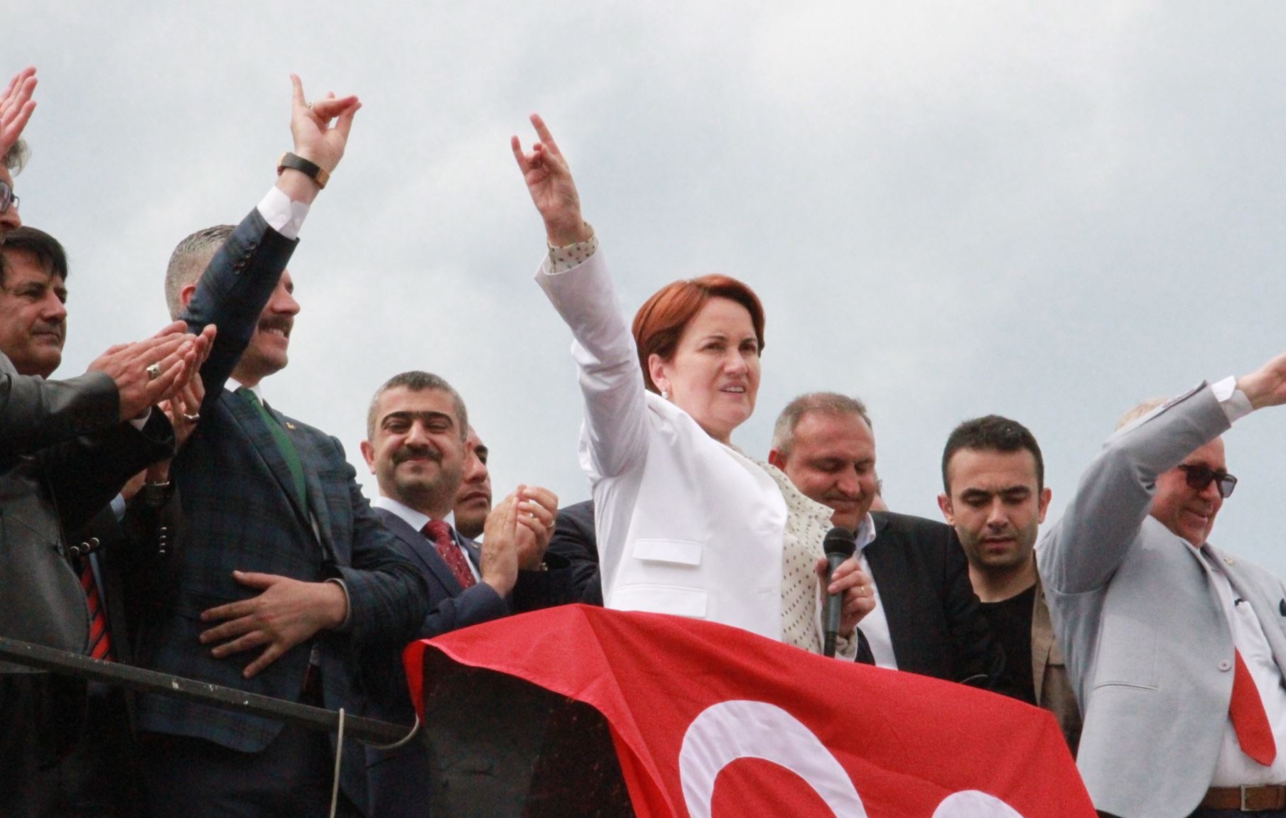 The image depicts a group of people at a political rally or event. In the foreground, a woman dressed in a white suit is speaking into a microphone, raising her arm with a gesture that signifies support or determination. Behind her, several men are visible, some clapping and others making similar gestures. The background features a large red banner, which may be associated with the event or political party. The overall atmosphere seems to convey enthusiasm and solidarity among the attendees.