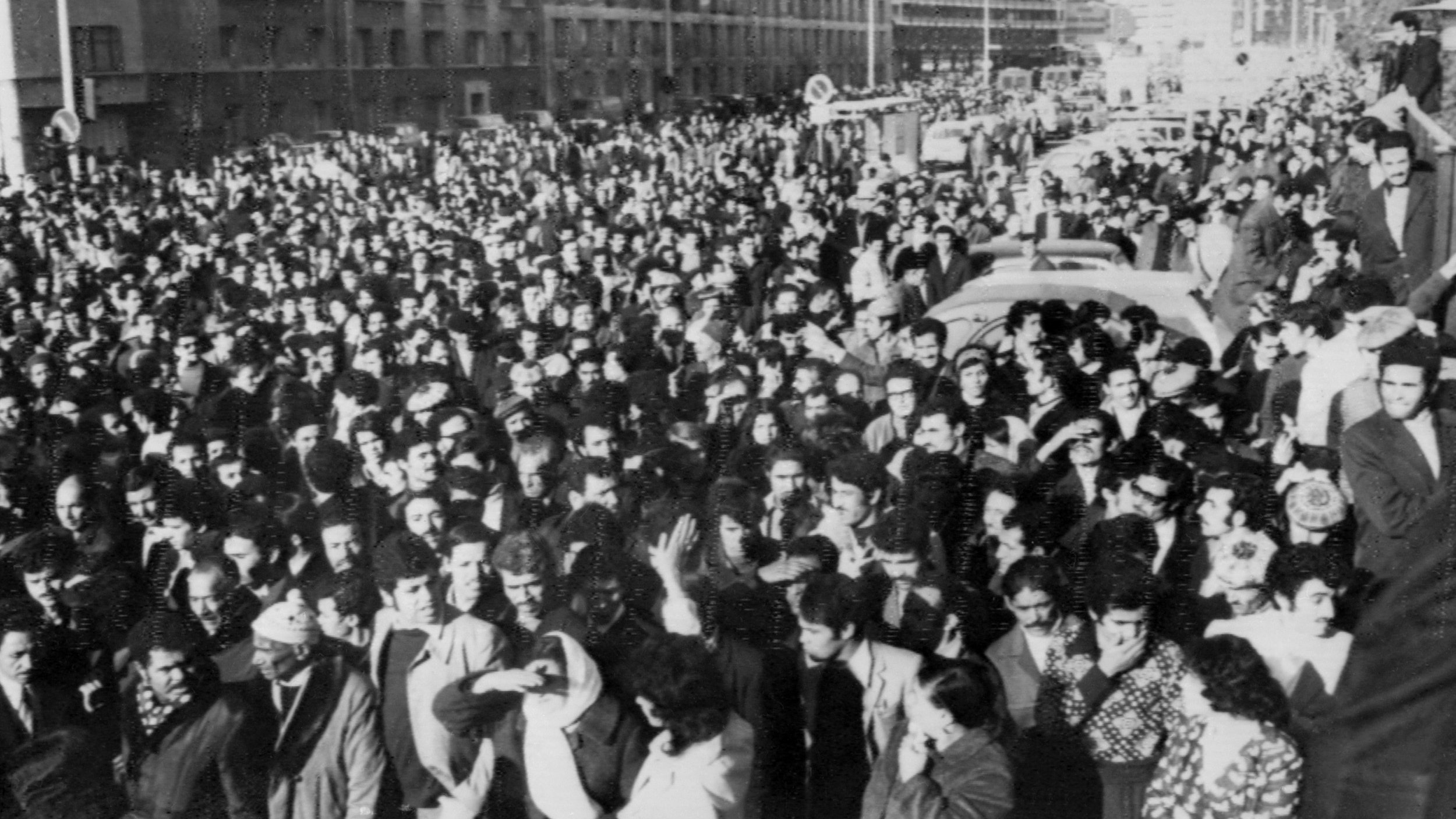 The image depicts a large crowd of people gathered in an urban setting, likely during a significant event or protest. The crowd appears to be densely packed, filling the street and surrounding areas. Many individuals are dressed in formal or semi-formal attire, indicating the seriousness of the occasion. The atmosphere seems charged, reflecting a sense of unity and purpose among the participants. Buildings line the background, and there are vehicles partially visible, suggesting a city environment. The black-and-white effect adds a historical or dramatic tone to the scene.