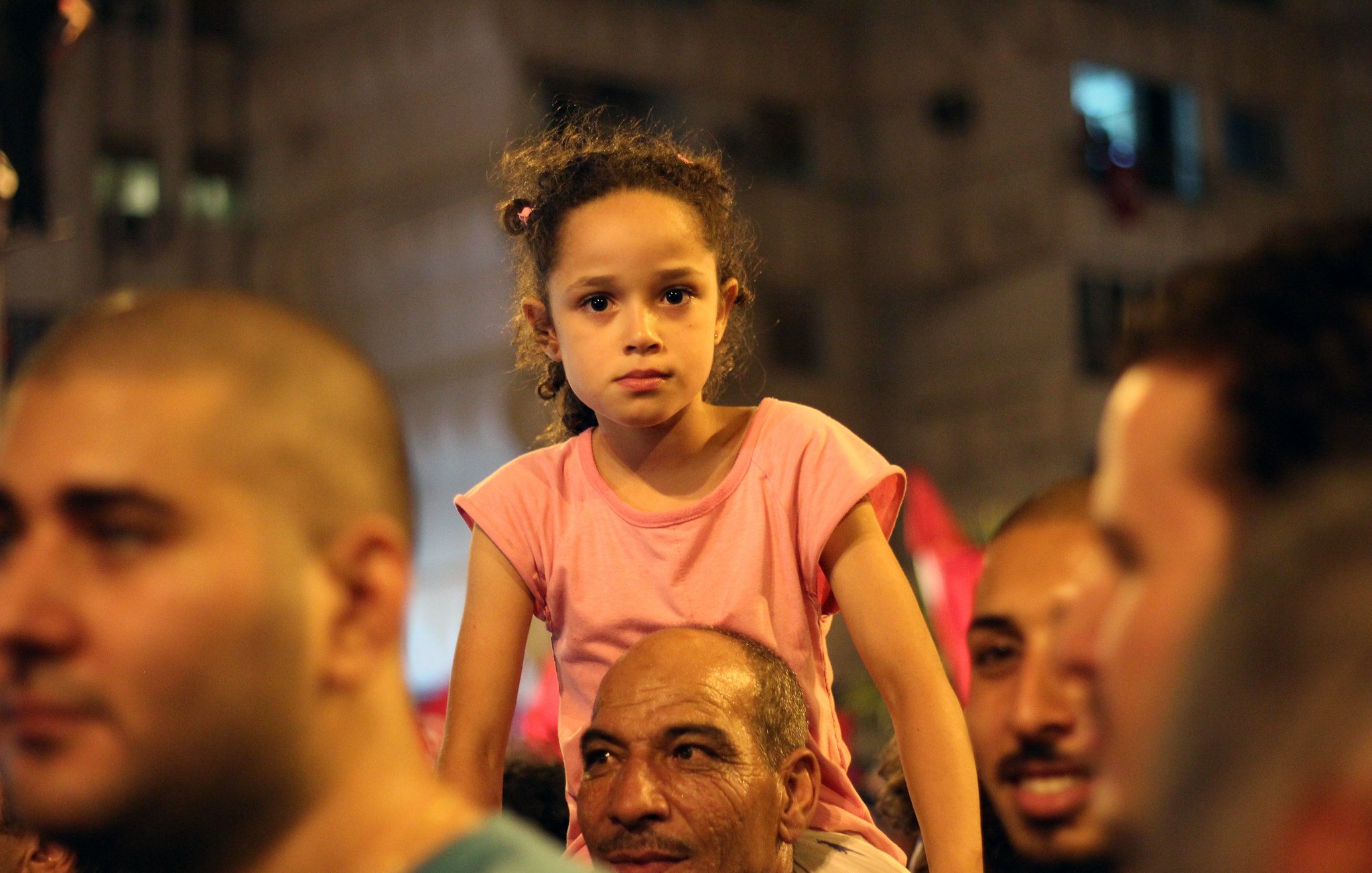 The image depicts a crowd at a public event or gathering. In the foreground, a little girl with curly hair, wearing a pink t-shirt, is seated on the shoulders of a man. She appears thoughtful or concerned, contrasting with the smiling faces of the adults around her. The background suggests a lively atmosphere, possibly filled with people and festive decorations. The scene captures a moment of reflection amid a bustling crowd.