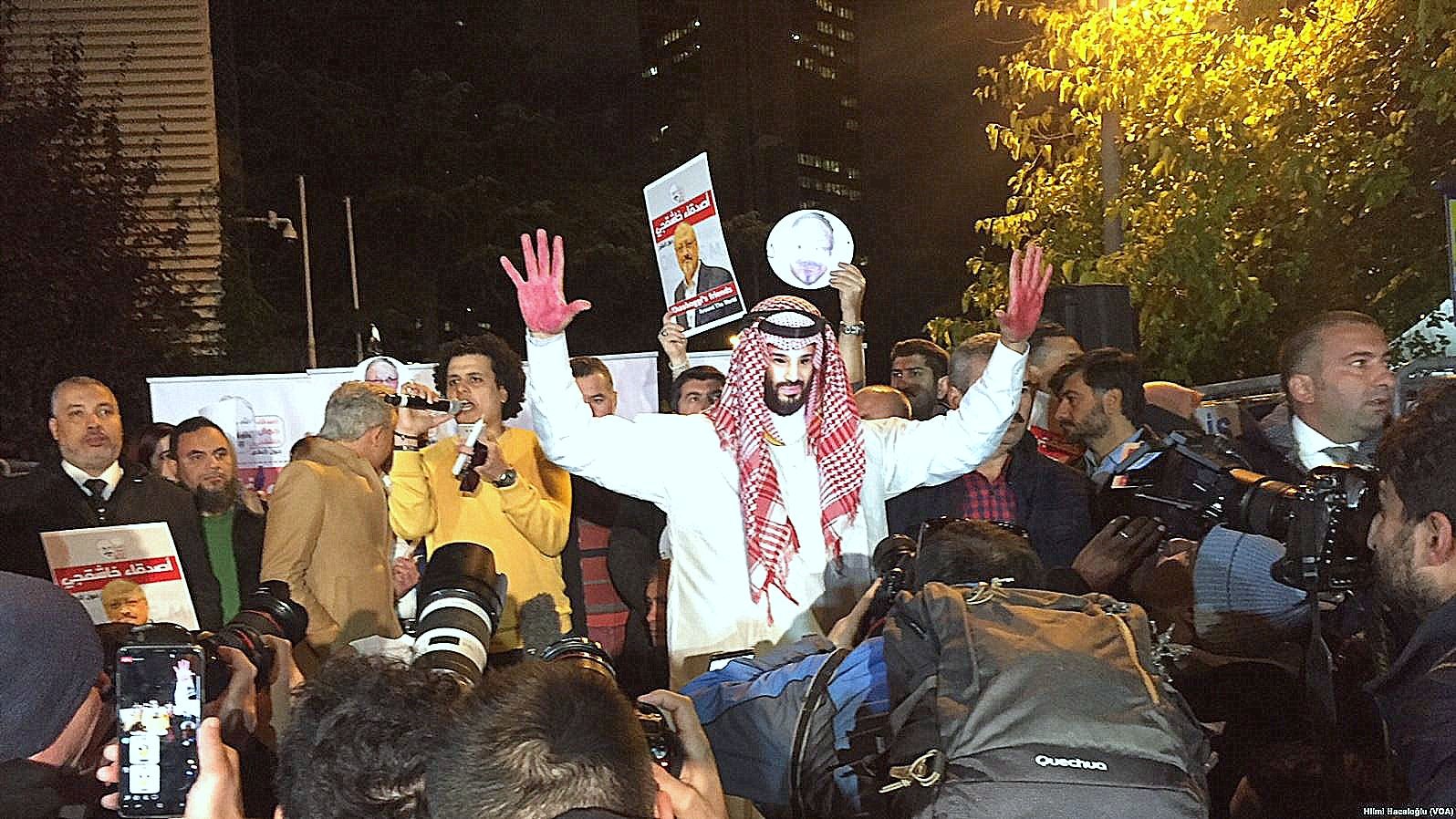 The image shows a group of people gathered for a protest or demonstration. In the foreground, a person wearing traditional attire, possibly a thobe and a headscarf, stands out. They have red paint on their hands, which could symbolize blood or a protest message. The individual is raising their hands and appears to be addressing the crowd or holding up a sign. Surrounding them are numerous cameras and reporters, indicating a media presence. The setting appears to be urban, with buildings visible in the background.
