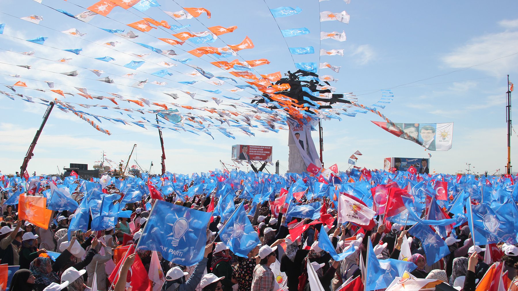 The image depicts a large gathering or rally, filled with a sea of people holding colorful flags. The flags appear to be in various shades of blue, orange, and other colors, creating a vibrant atmosphere. Many attendees are wearing white hats and there are banners or signs visible in the background. The sky is bright, suggesting a sunny day, and the scene conveys a sense of energy and excitement among the crowd.