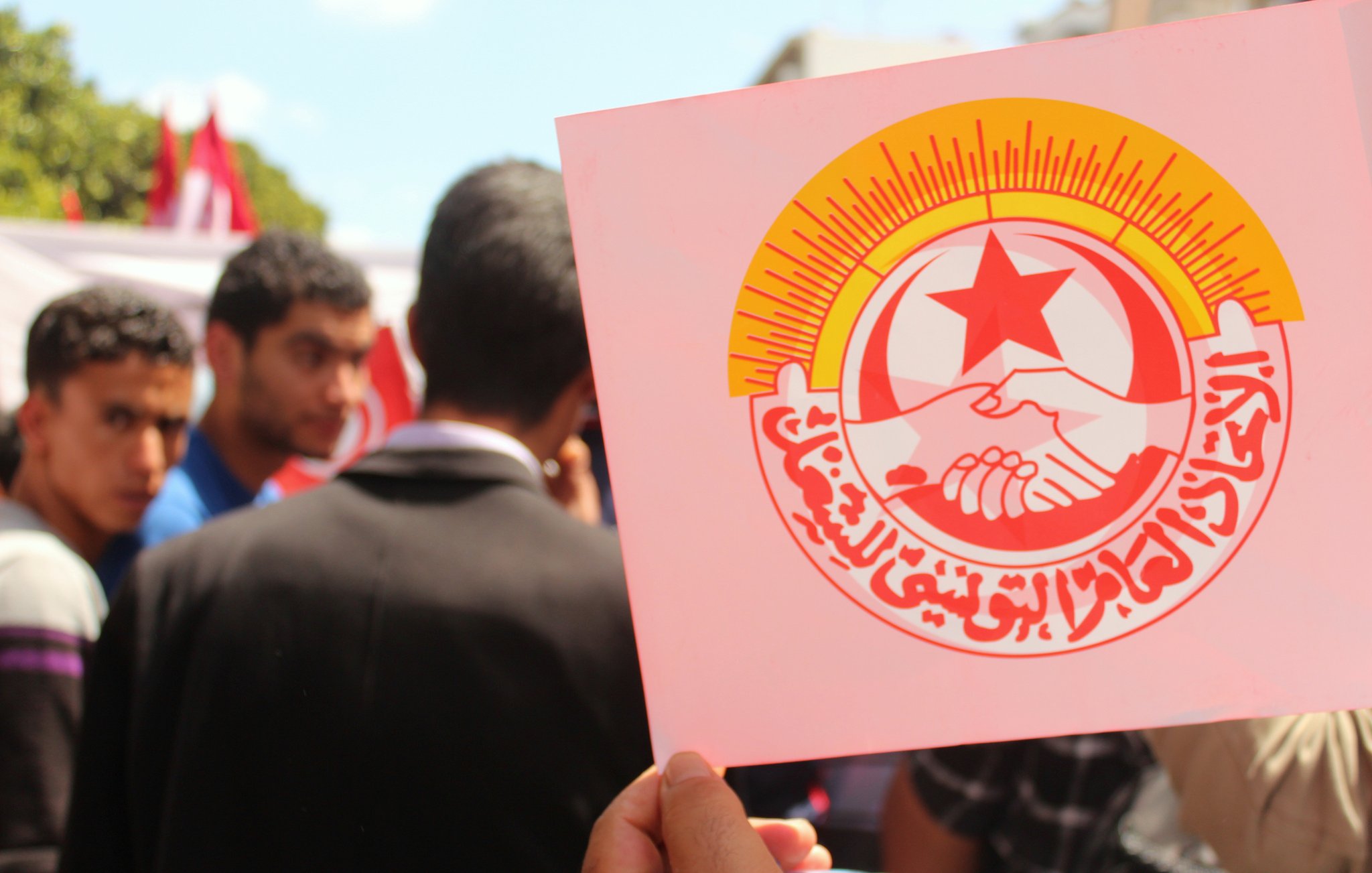 The image features a group of people, with a focus on one individual holding up a sign. The sign prominently displays a logo that includes two hands shaking, surrounded by a bright sunburst and red star. The text on the sign is in Arabic, likely associated with a labor or union organization. The background suggests an outdoor setting, possibly during a rally or demonstration, with other individuals visible nearby.