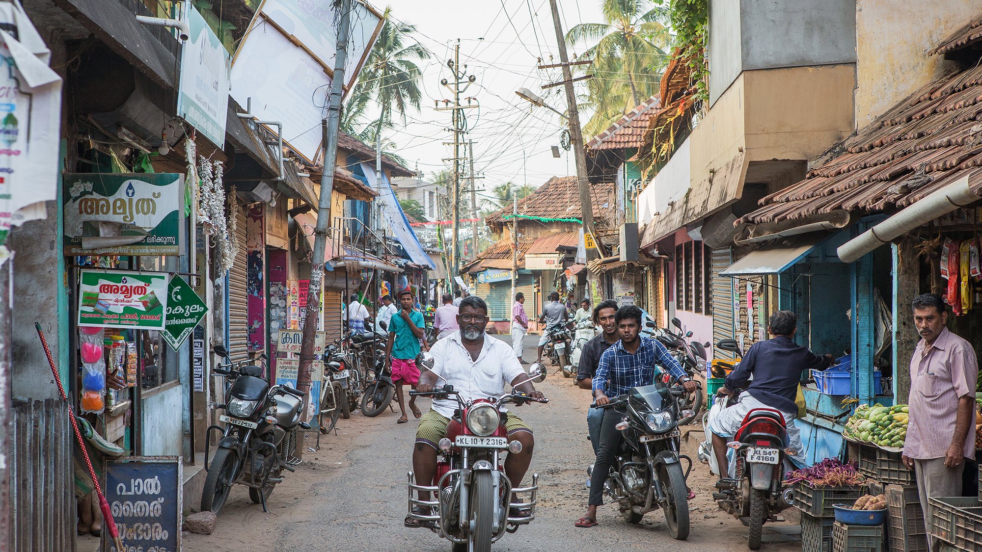 The image depicts a bustling street scene, likely in a South Asian locale. There are narrow, lined pathways with a mixture of residential and commercial buildings, featuring traditional architecture. People can be seen engaging in everyday activities, some on foot and others riding motorcycles. Shops are displaying various goods, and there are signs in a local language. The atmosphere appears lively, with palm trees in the background enhancing the tropical feel of the setting.