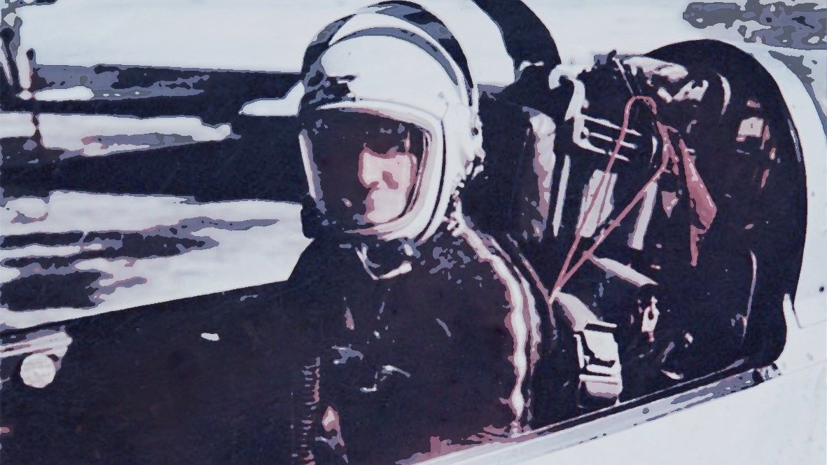The image depicts a person seated in a cockpit, likely a pilot or astronaut. They are wearing a helmet and protective gear, suggesting they are preparing for a flight or mission. The background appears somewhat blurred, emphasizing the individual in the cockpit. The overall tone of the image conveys a sense of readiness and focus, typical of aviation or space exploration contexts.