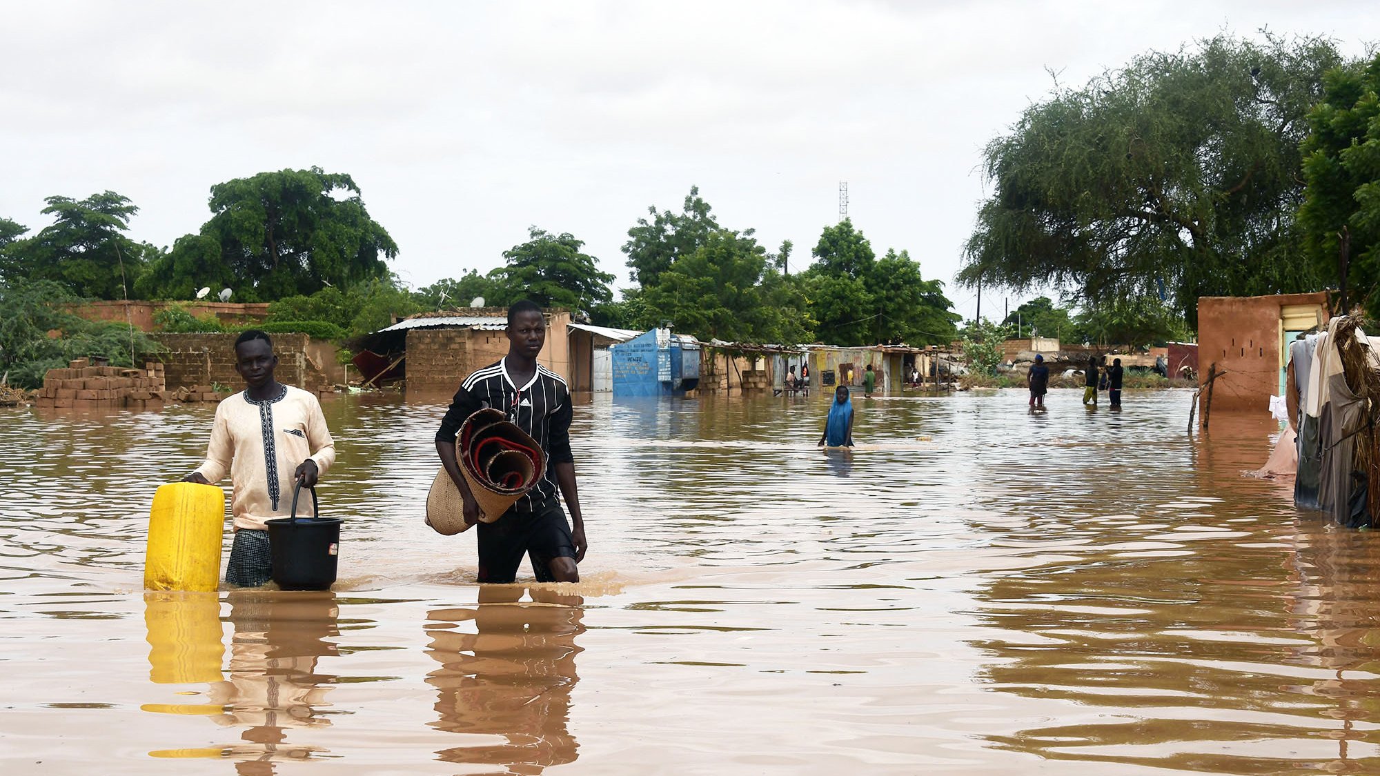The image depicts a flooded area, where individuals are walking through standing water. Two men are central in the foreground; one is carrying a bucket, while the other holds a rolled-up mat. In the background, makeshift shelters or homes are partially submerged, surrounded by trees and greenery. The scene reflects the impact of flooding, with water covering the ground and affecting the surrounding environment. The sky is overcast, suggesting ongoing adverse weather conditions.