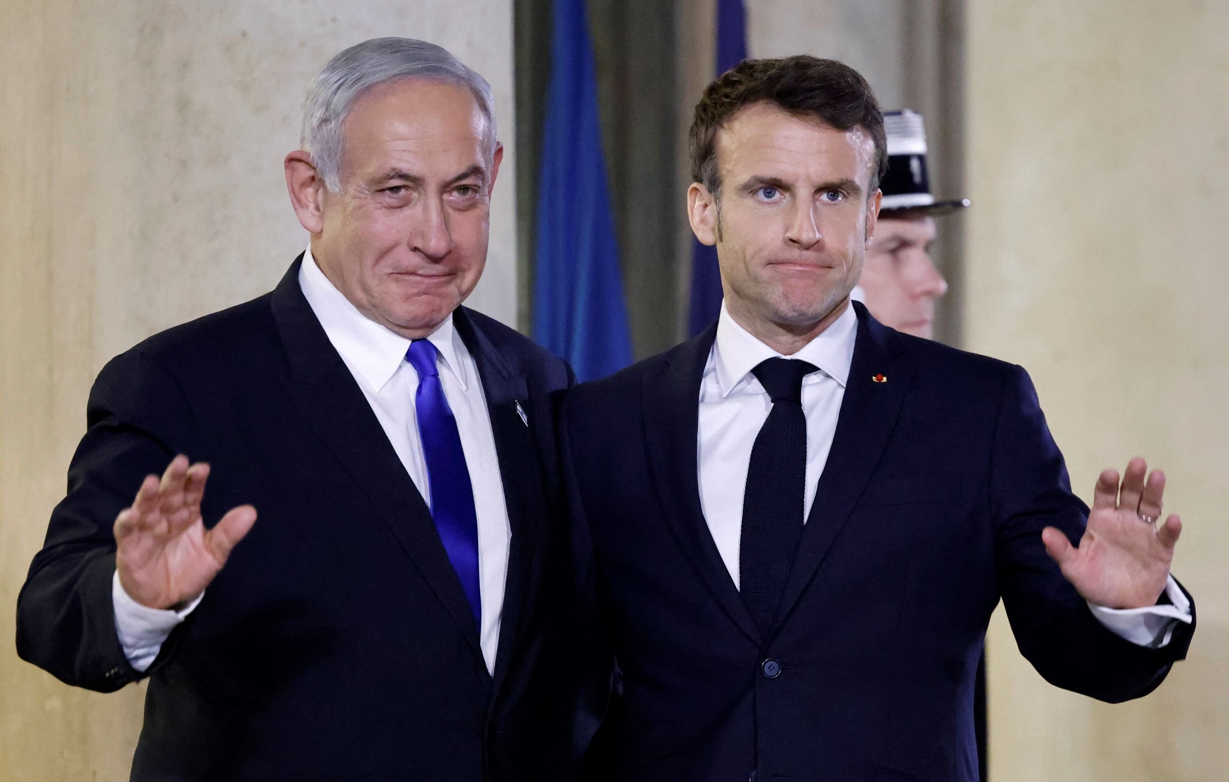 The image shows two men standing side by side, both dressed in formal attire. One is wearing a blue tie and the other has a dark tie, indicating a formal event. They appear to be waving, possibly to an audience or camera. The background suggests an official setting, likely an indoor venue with flags or formal decorations. Their expressions seem to convey a mix of seriousness and cordiality.