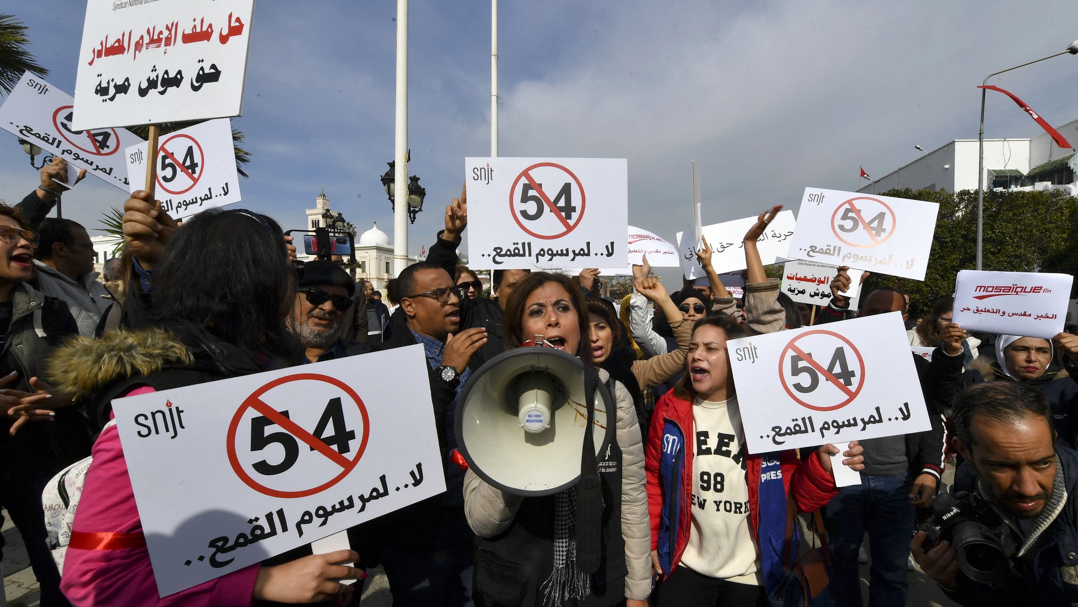 The image depicts a protest scene with a group of people holding up signs and banners. Some demonstrators are prominently featuring a sign with the number "54" crossed out, suggesting opposition to a specific law or regulation. One woman is speaking into a megaphone, leading the crowd in their protest. The atmosphere appears energetic and determined, with a focus on raising awareness or voicing their concerns regarding a particular issue.