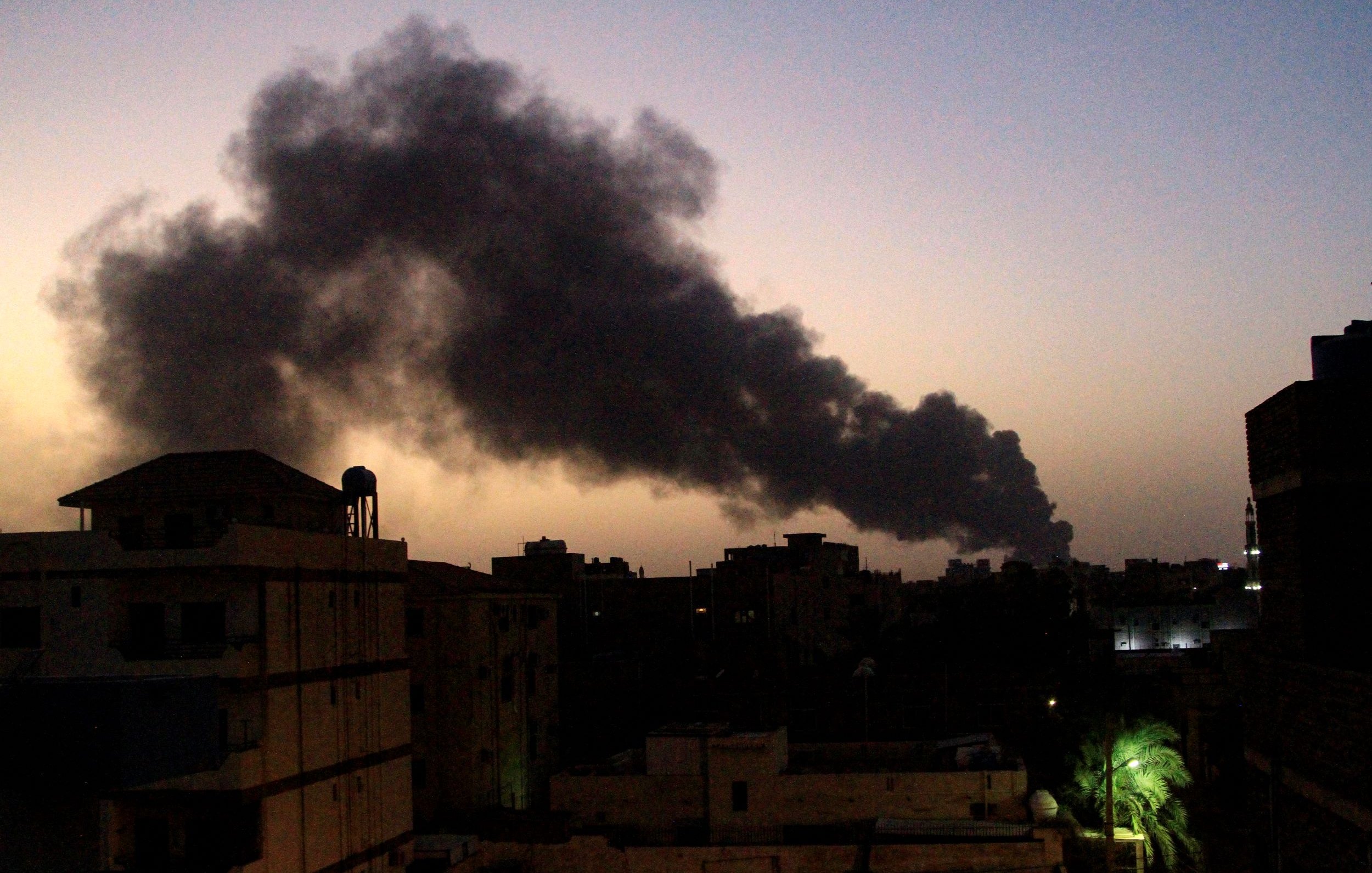 The image depicts a dusk or evening scene in an urban environment, with several buildings visible in the foreground. In the background, a substantial plume of black smoke rises into the sky, creating a stark contrast against the fading light of sunset. The smoke suggests a fire or some kind of industrial activity. The overall mood of the image conveys a sense of urgency or concern, given the presence of the smoke.