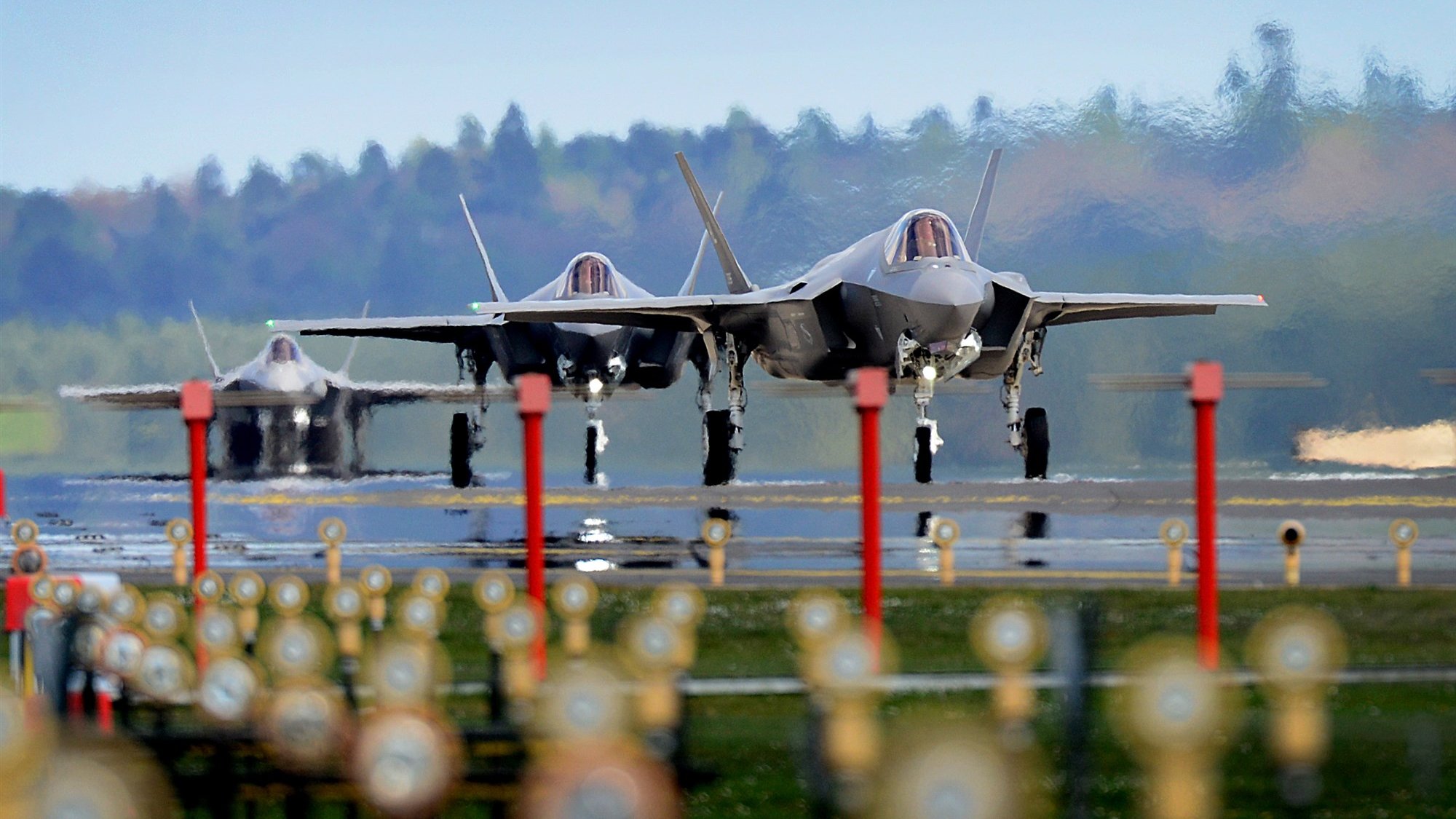 The image features three F-35 fighter jets on an airstrip, showcasing their sleek and modern designs. The jets are positioned closely together, and the background features a blurred green landscape, indicating a rural setting. The foreground includes several lights lining the runway, which adds a sense of depth to the scene. The overall atmosphere appears dynamic and focused on military aviation.