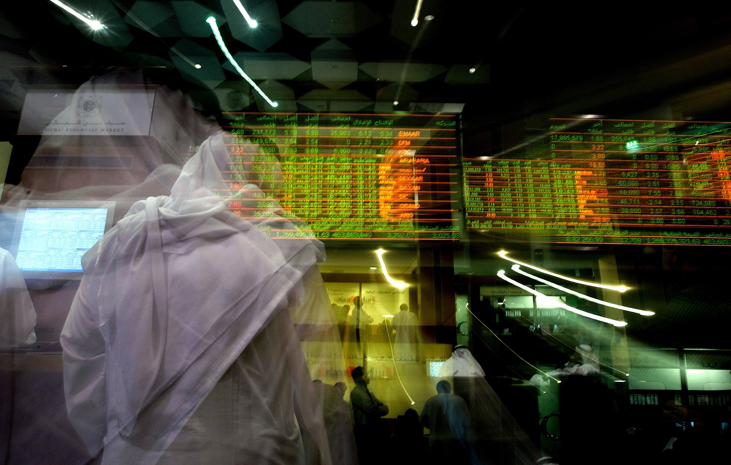 The image depicts a bustling financial trading environment, likely in a stock exchange. There are dynamic digital displays showing stock prices and market information, with green and red indicators reflecting changes in values. The foreground features figures in traditional attire, possibly indicating a cultural context. The overall scene conveys a sense of movement and activity, highlighting the fast-paced nature of trading. The use of motion blur adds to the dynamic feel of the environment.