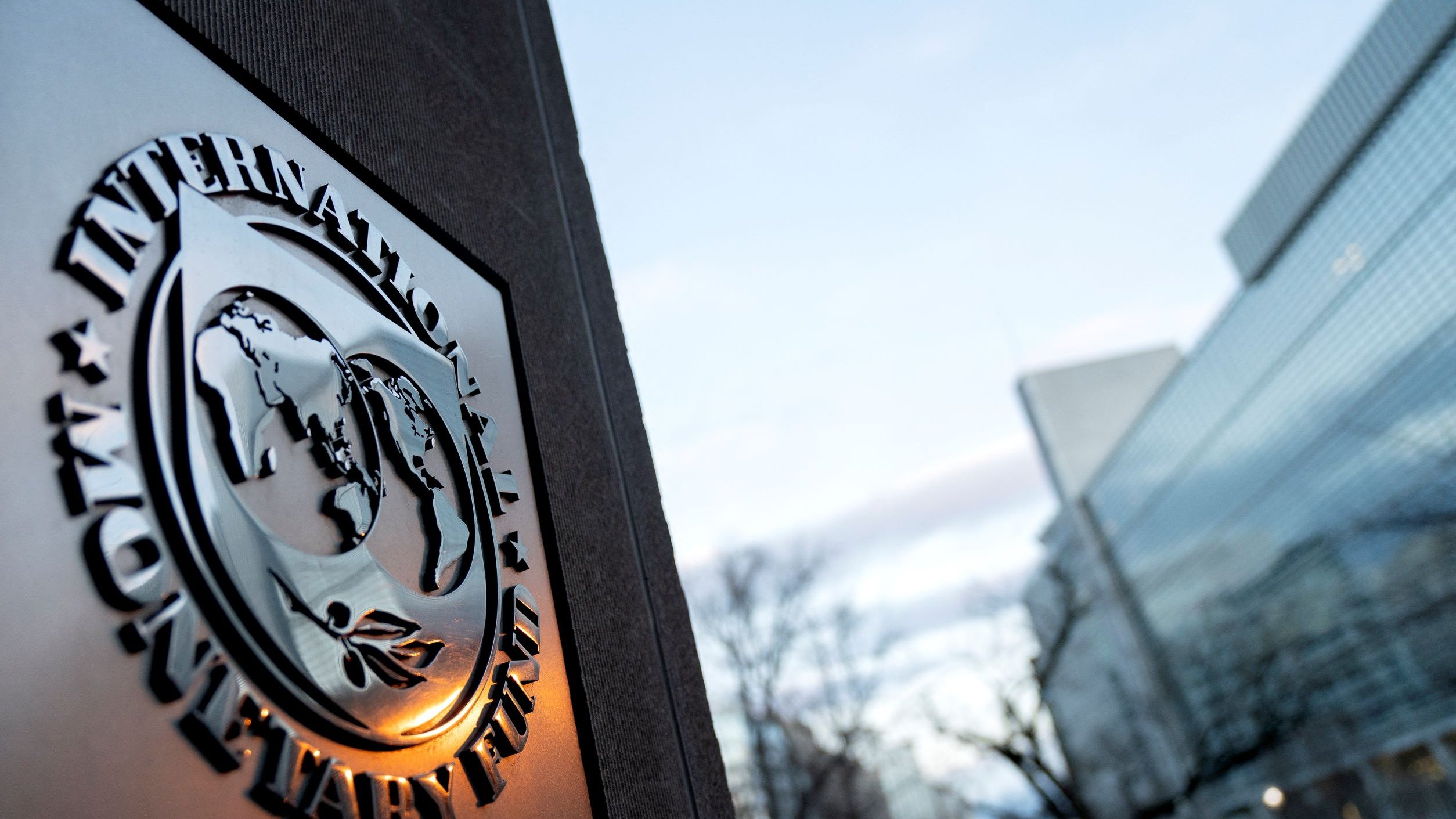 The image features the emblem of the International Monetary Fund (IMF) prominently displayed on a stone surface. The emblem includes a globe surrounded by olive branches, symbolizing peace and cooperation. In the background, there is a modern building with glass windows, reflecting a serene sky with some clouds. The setting appears to be urban and professional, likely outside the IMF headquarters.