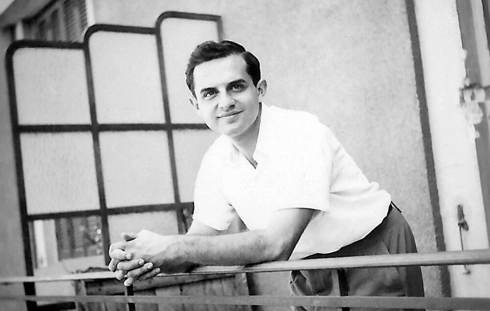 The image features a young man with a pleasant expression, leaning on a railing. He has dark hair styled in a neat manner and is dressed in a light-colored short-sleeve shirt paired with trousers. The background suggests a balcony or outdoor setting, with a partially visible modern-style partition or screen. The photo has a vintage quality, indicating it was likely taken several decades ago.