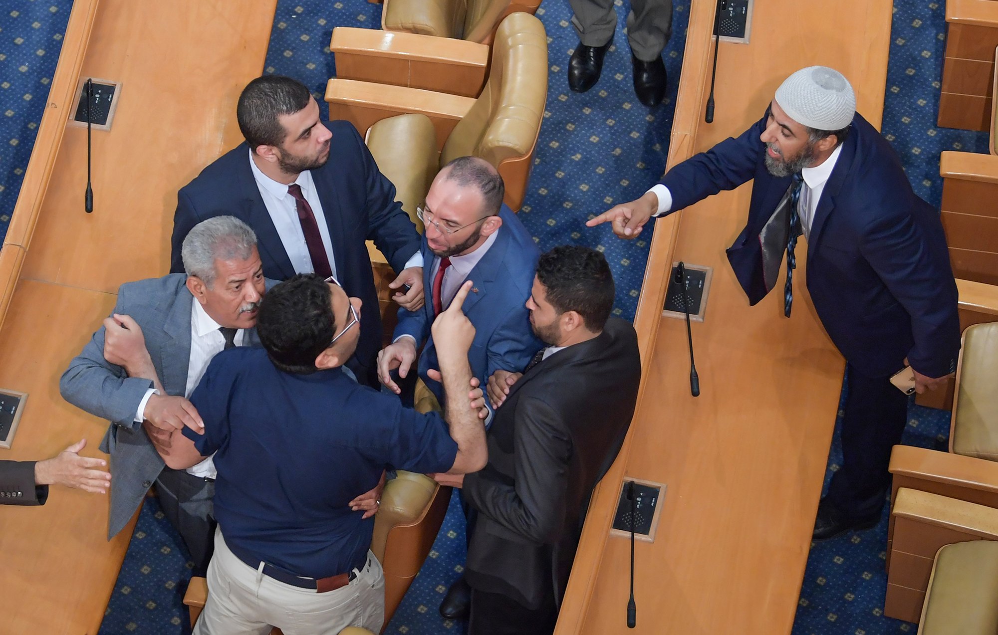 The image shows a heated confrontation occurring in a legislative assembly. Several men are engaged in a tense discussion or argument, with some gesturing animatedly. The setting appears to be a formal chamber, indicated by the rows of seats and the official appearance of the individuals. Some men are standing closely together, while others seem to be pointing or raising their hands, indicating strong emotions. Overall, the scene conveys a moment of conflict or debate among the legislators.