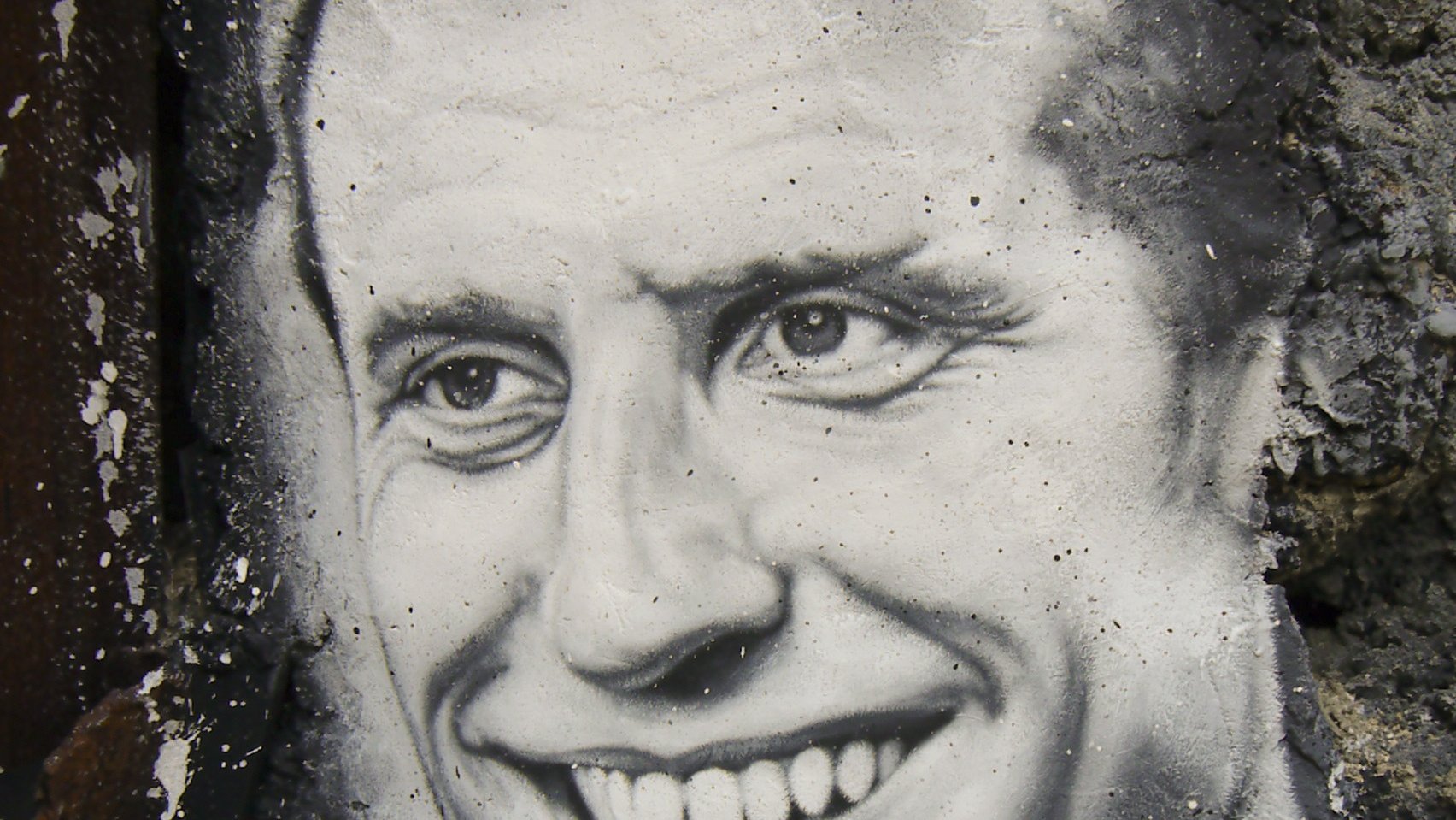 The image features a black-and-white mural of a smiling man. The artwork is set against a textured, stone wall, which adds depth to the scene. The man's expression conveys warmth and friendliness, with a prominent smile and bright eyes. The details are finely rendered, showcasing the artist's skill in capturing facial features and expressions.