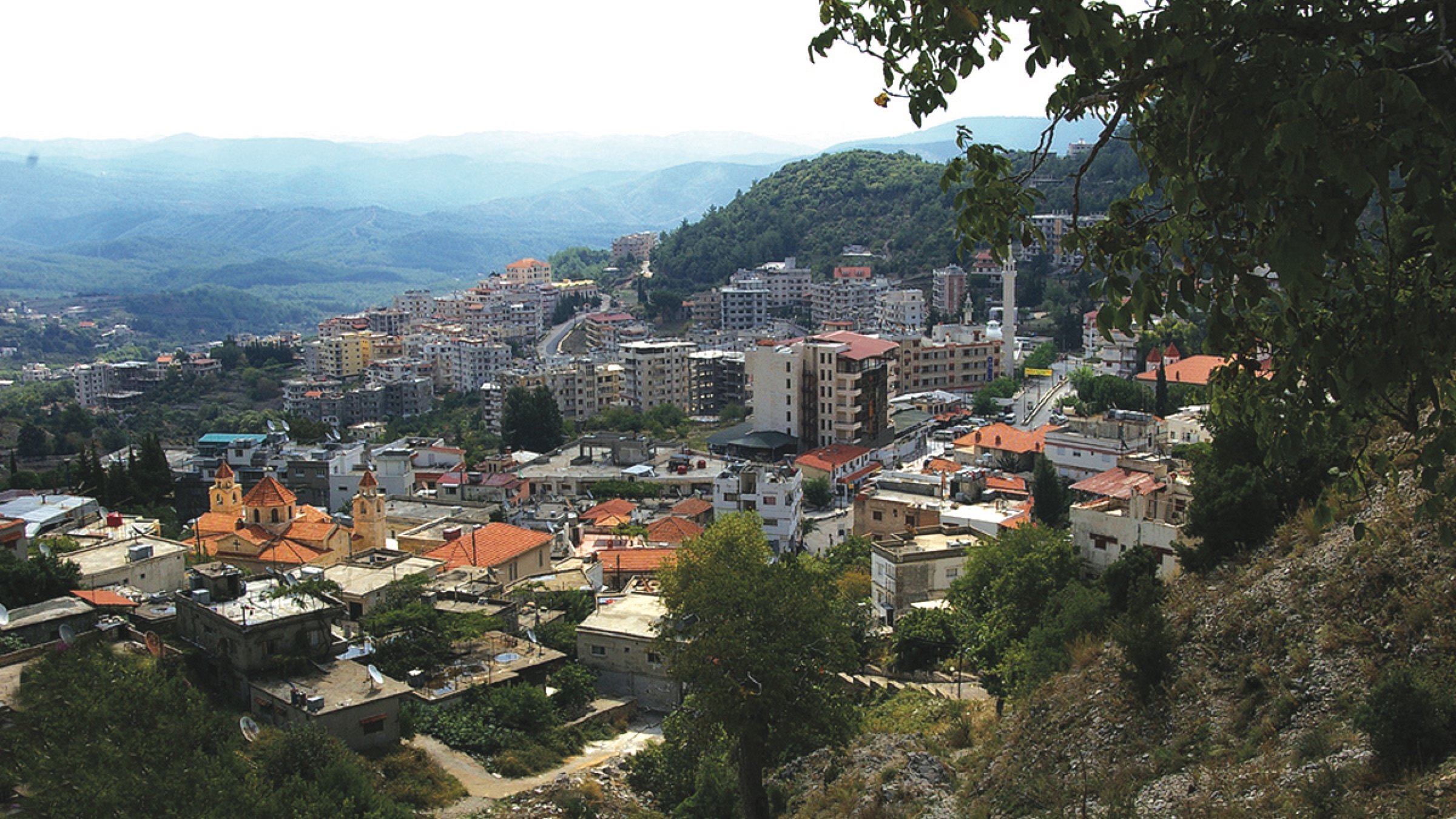 The image shows a panoramic view of a mountainous landscape with a town nestled in the valleys. The foreground features a mix of low-rise buildings, some with red rooftops, and greenery, including trees and hills. In the background, a larger urban area can be seen with taller buildings, likely indicating a more developed part of the town. The sky appears slightly overcast, with mountains extending into the distance, adding depth to the scene. The overall atmosphere suggests a blend of natural beauty and urban development.