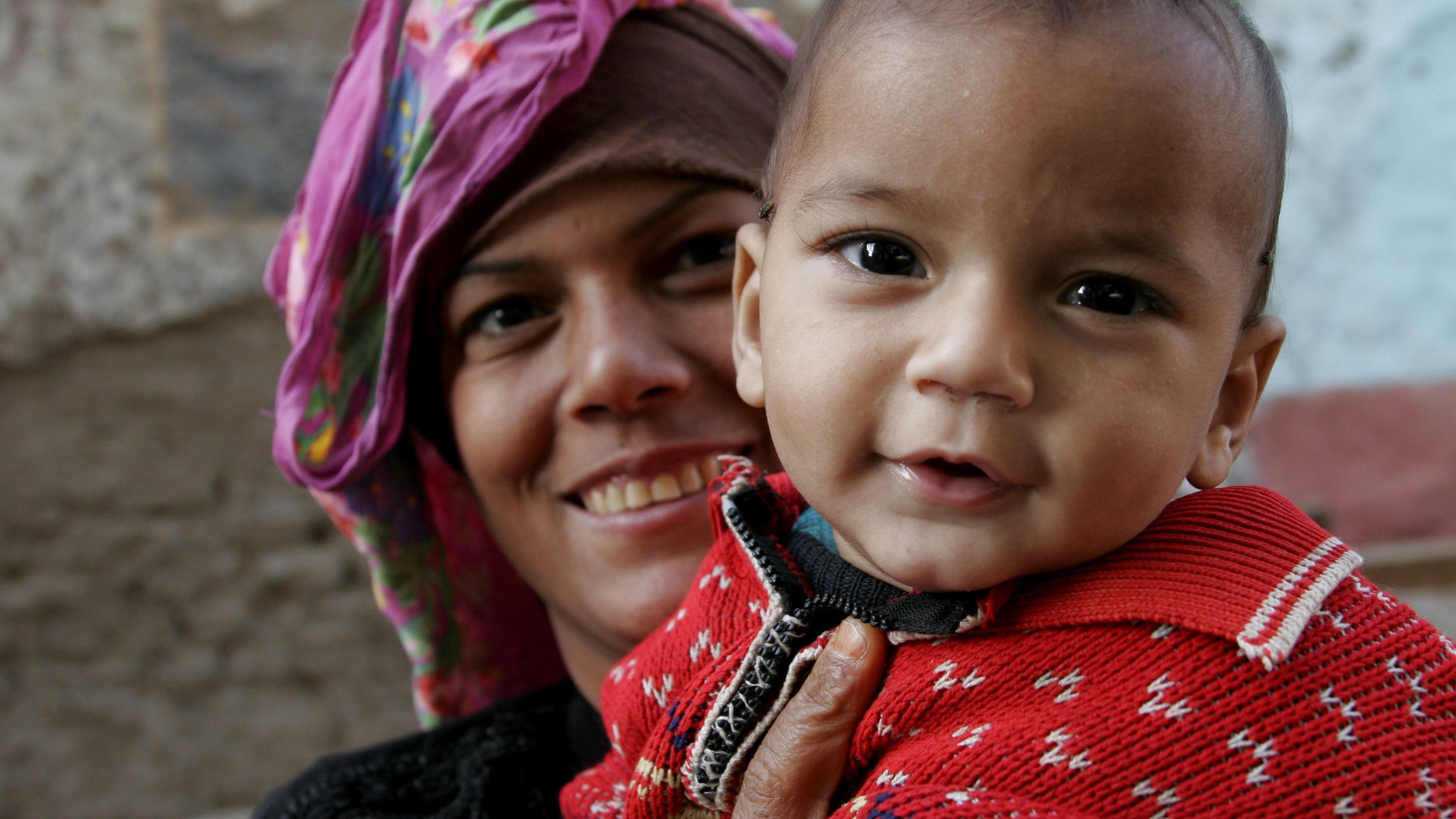 The image features a smiling woman holding a small child. The woman is wearing a colorful headscarf, and her expression is warm and cheerful. The child, who is dressed in a bright red sweater with a patterned design, looks happy and is facing the camera, showing a playful smile. The background appears to be a textured wall, suggesting an outdoor or rustic setting. The overall mood of the image is joyful and intimate, capturing a moment of connection between the woman and the child.