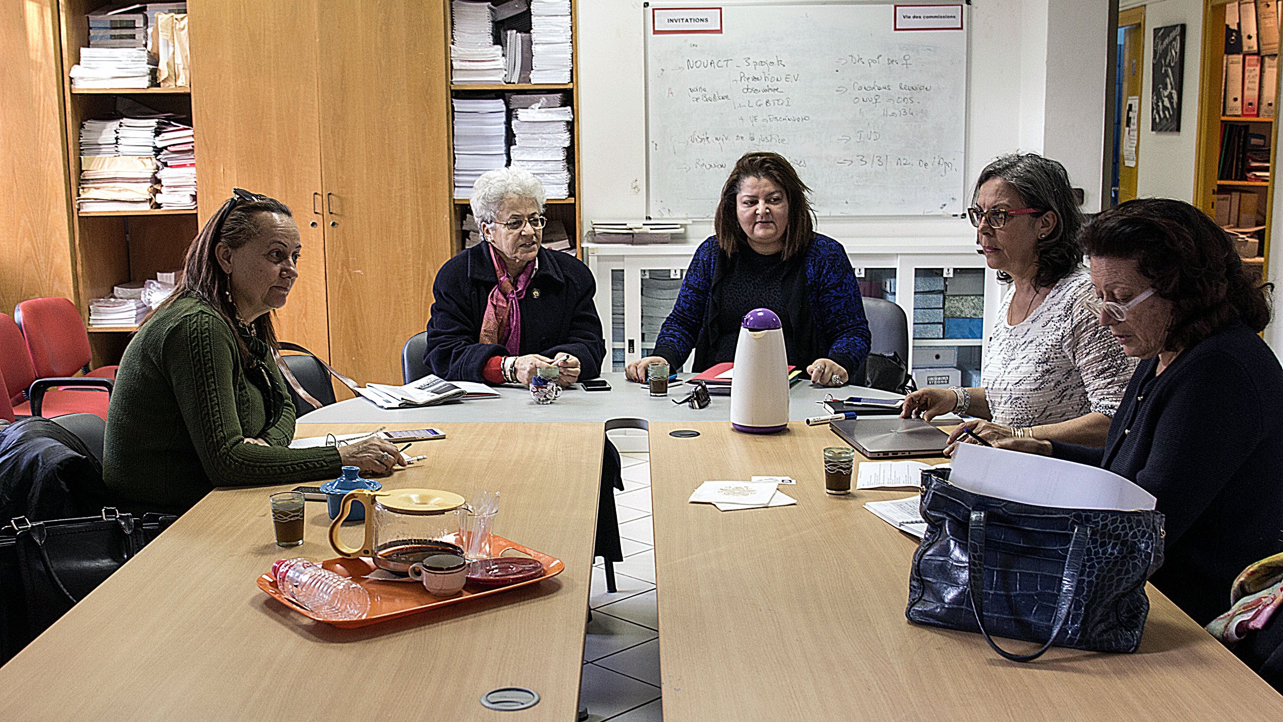 The image depicts a group of five women sitting around a table in a room filled with shelves and papers. They appear to be engaged in a discussion or work-related activity. The table has some items on it, including drinks and papers, suggesting a collaborative atmosphere. The women are looking at each other, indicating communication and interaction among them. The setting seems to be an office or a meeting room.