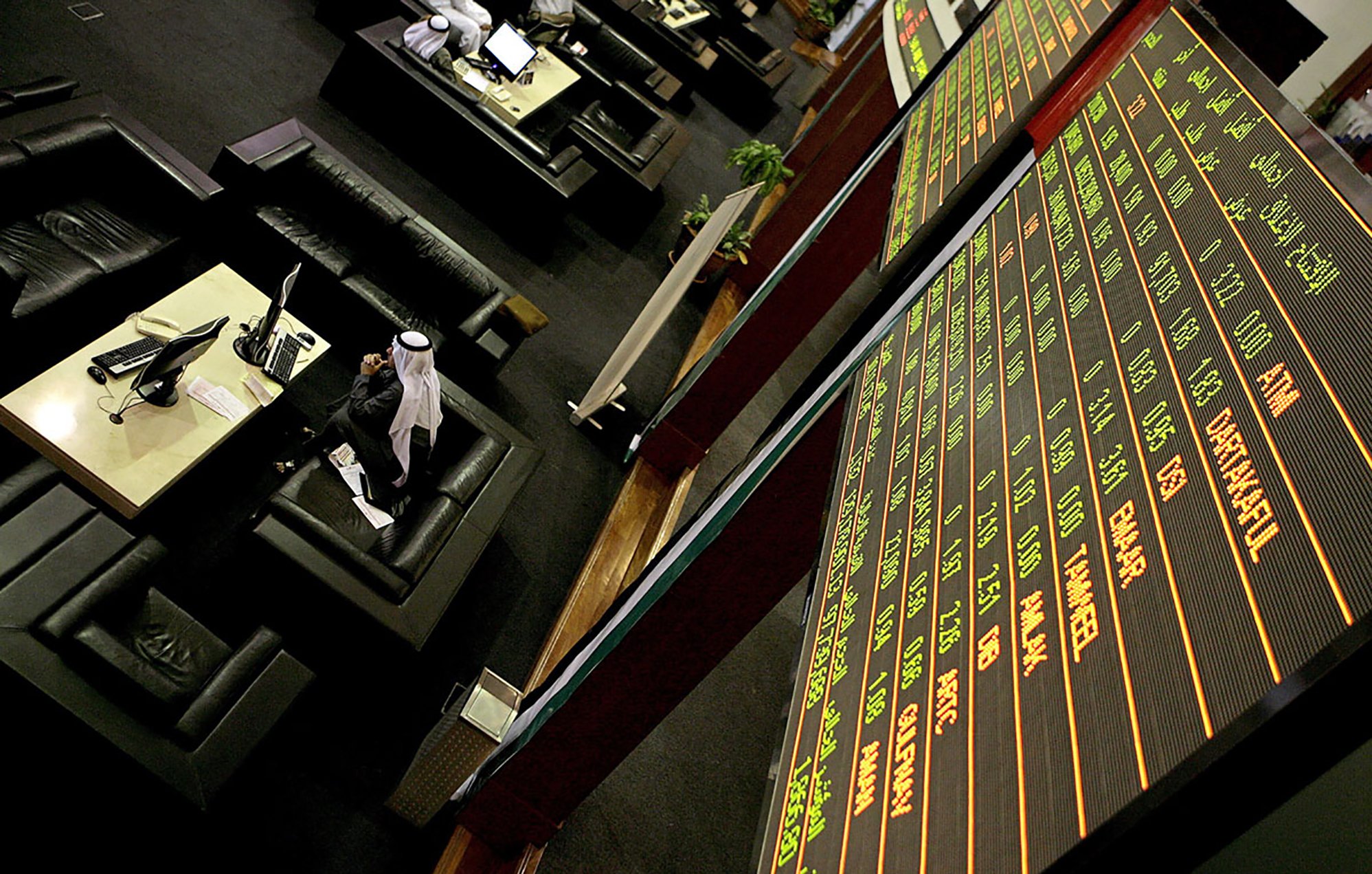 The image depicts a financial trading environment, likely within a stock exchange. In the foreground, there is a large electronic display board showing various financial data, such as stock prices and indices, with green and red text indicating changes in value. Below the board, there are several black leather seating areas where traders or visitors might be sitting. One individual, dressed in traditional attire, appears to be engaged with a computer or taking notes. The overall atmosphere suggests a busy and professional financial setting.