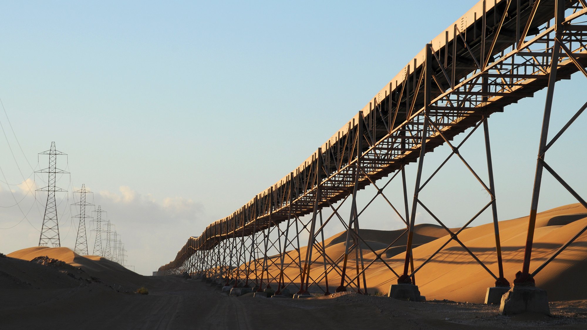 The image depicts a long conveyor belt system stretching across a vast landscape dominated by sand dunes. The conveyor belt is elevated on metal supports, running parallel to the ground. In the background, tall power lines extend into the horizon, hinting at an industrial setting. The warm light of the sun creates a golden hue on the sand, adding to the serene yet striking composition of the scene. The overall atmosphere suggests a remote, arid environment that may be associated with mining or agricultural operations.