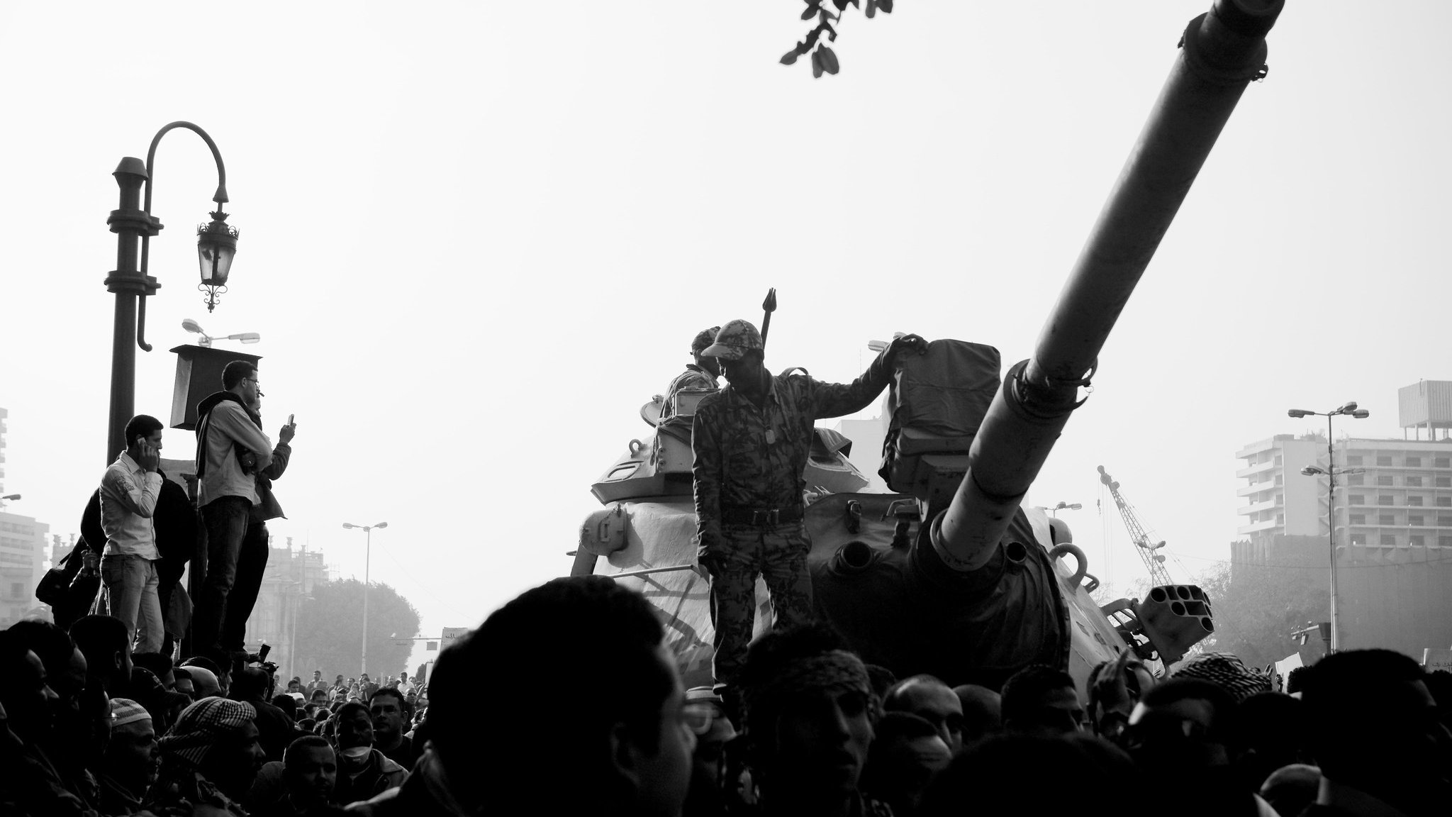 The image depicts a scene of intense activity, likely in an urban setting. In the foreground, a soldier stands on a tank, raising an arm, possibly in a gesture of authority or encouragement. The background is filled with a dense crowd, indicating a significant public event, possibly a protest or military presence. The overall tone of the image is stark, being presented in black and white, which adds to its dramatic effect. The atmosphere seems charged with emotion and energy, with some individuals observing or participating actively in the scene.